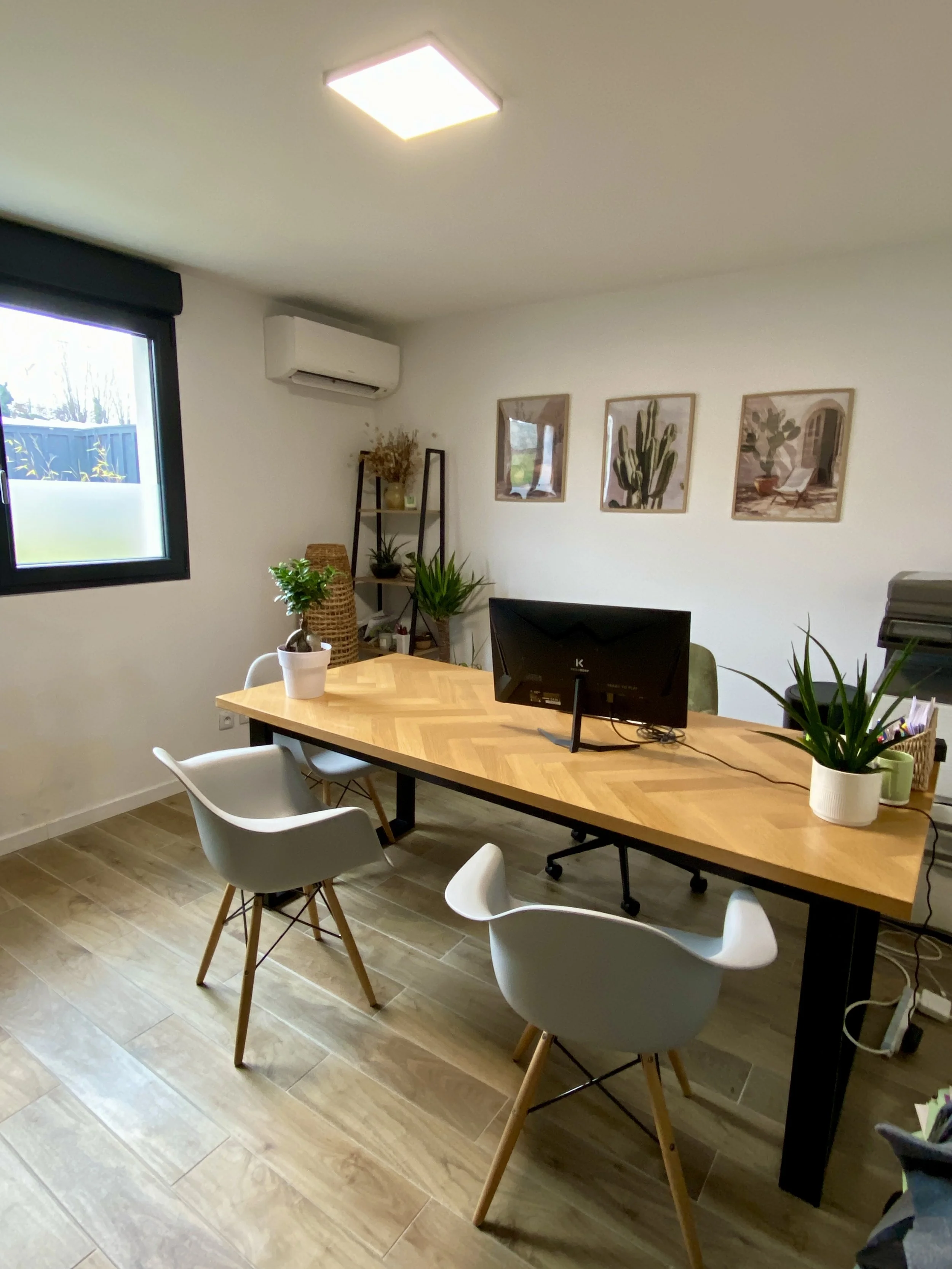 Bureau avec un bureau en bois, trois chaises blanches, plantes en pot, une fenêtre, et des cadres de décoration sur le mur.