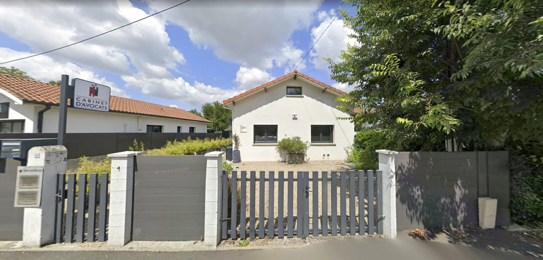 Maison blanche avec un toit en tuiles rouges, jardin avec des plantes, clôture grise, signale un cabinet d'avocats.