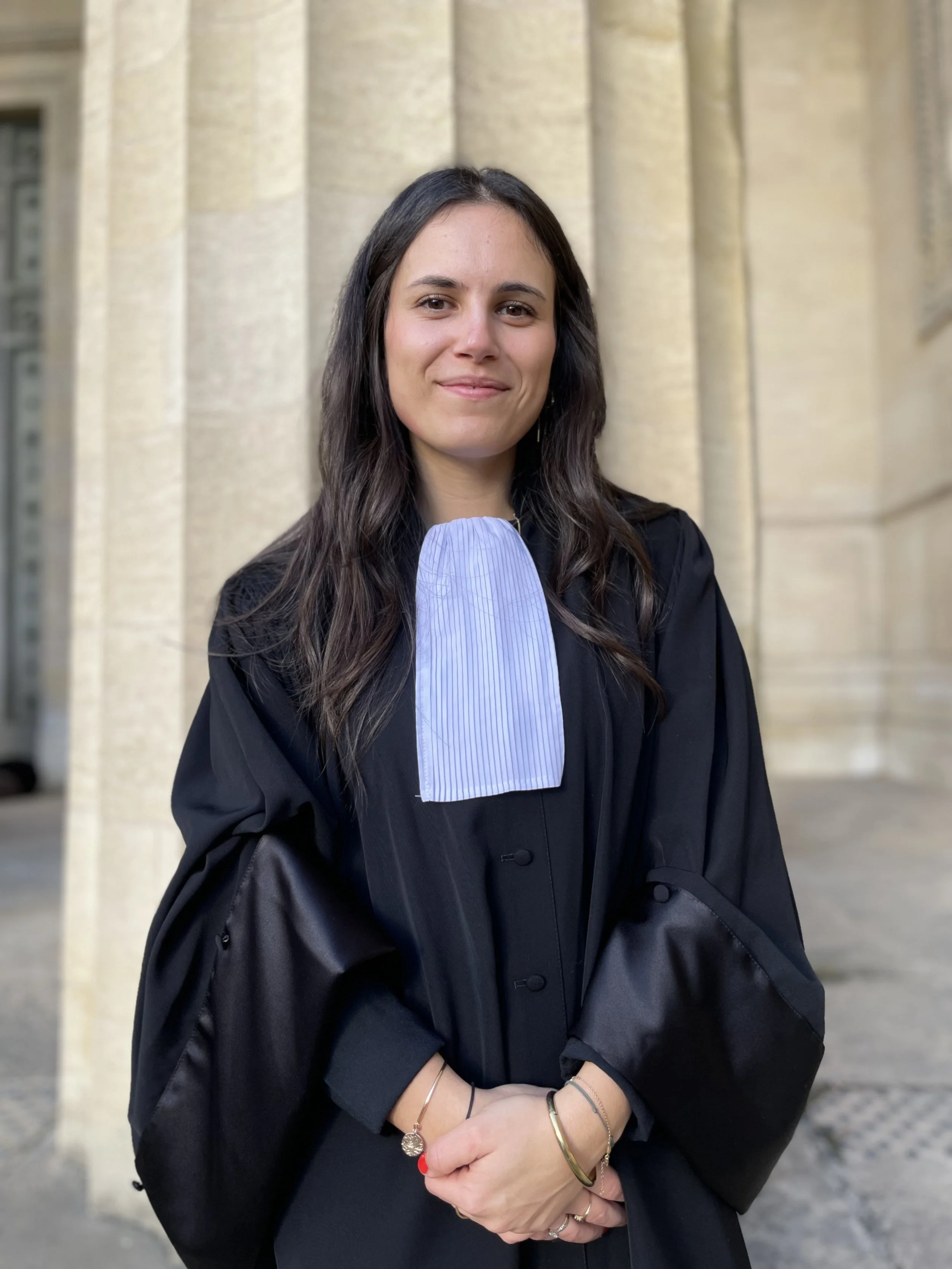 Une femme en robe d'avocat aux couleurs sombres avec un collier blanc à rayures bleues, posant devant un bâtiment en pierre, probablement une cour de justice.