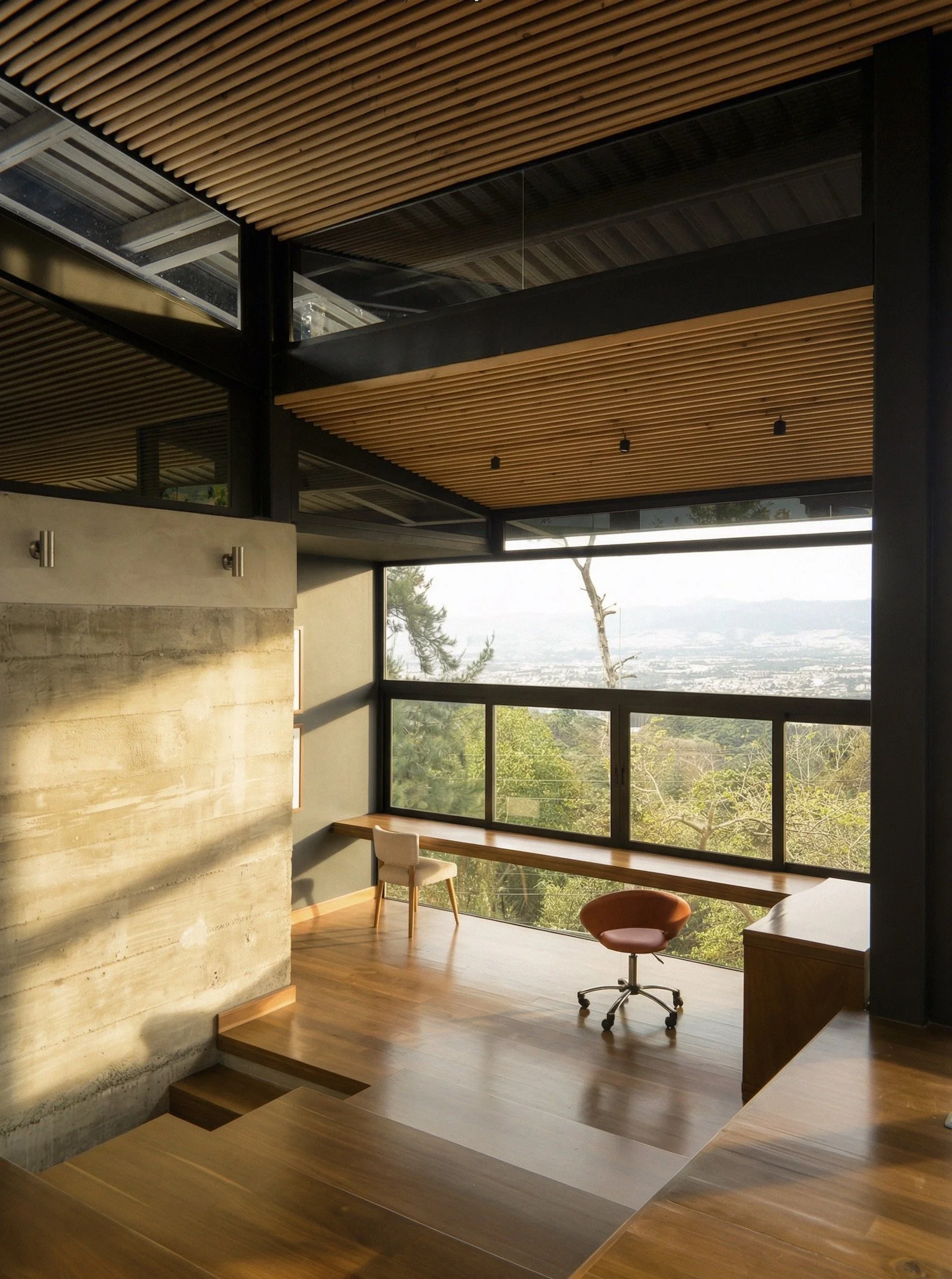Modern interior room with large windows and a view of trees and cityscape, featuring a wooden ceiling, a concrete wall, and minimal furniture including a desk and chairs.