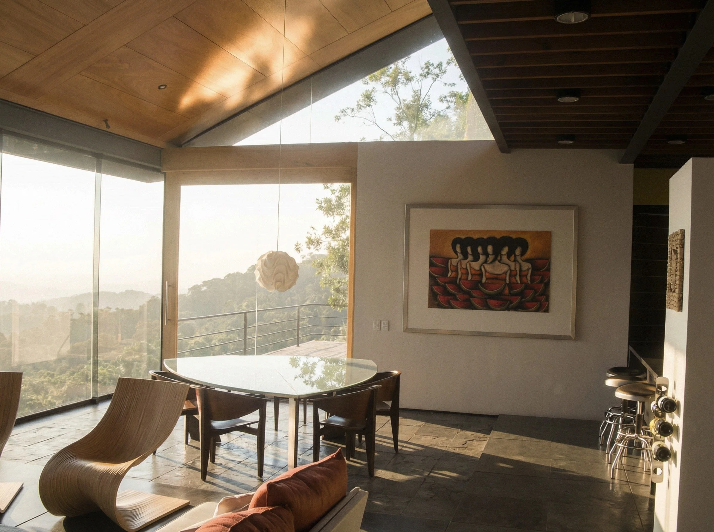 Modern dining area with a glass top table, surrounded by wooden chairs, in a room with large windows and a mountain view, decorated with abstract art and a sculptural pendant light.