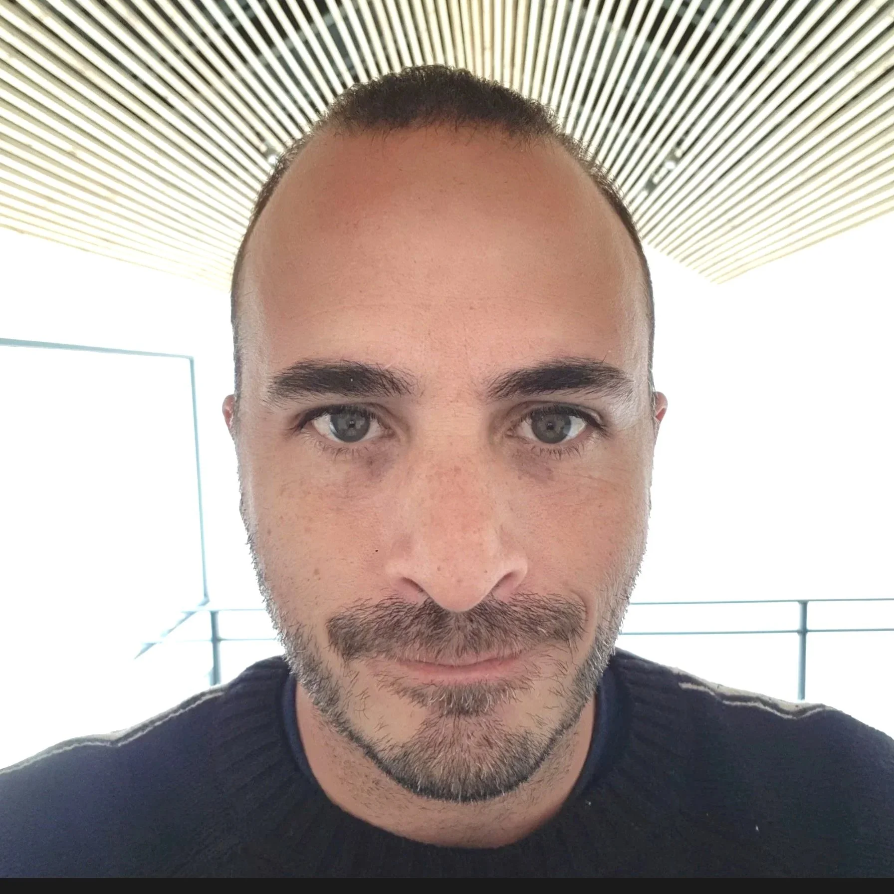 Close-up of a man with short dark hair, blue eyes, and a beard, wearing a black shirt, indoors with a wooden slatted ceiling and white background.