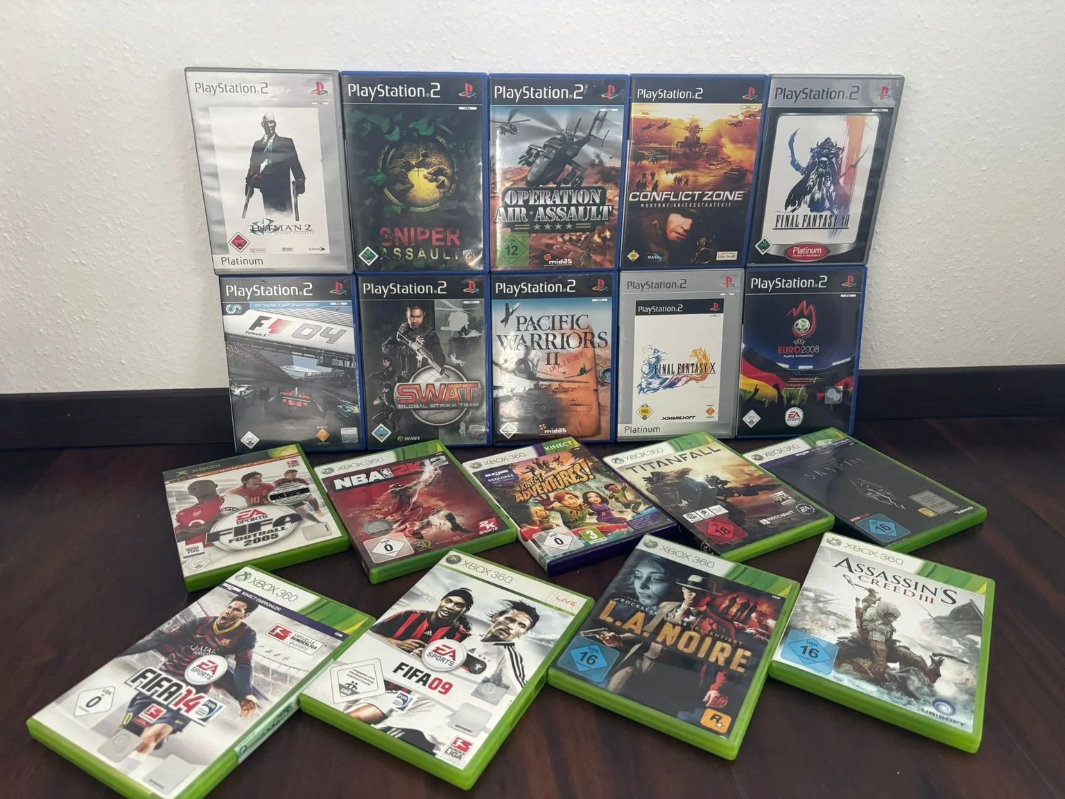Mehrere Spieledosen und -hüllen für Konsolen auf einem Tisch, darunter PlayStation 2 und Xbox 360 Spiele wie 'Hitman 2', 'Sniper Assault', 'Operation Air Assault', 'Conflict Zone', 'Final Fantasy', 'F1 04', 'SWAT', 'Pacific Warriors', 'Final Fantasy X', 'Euro 2008', 'NBA 2K5', 'Kinect Adventures!', 'Titanfall', 'Star Wars', 'FIFA 14', 'FIFA 09' und 'Assassin's Creed II'.