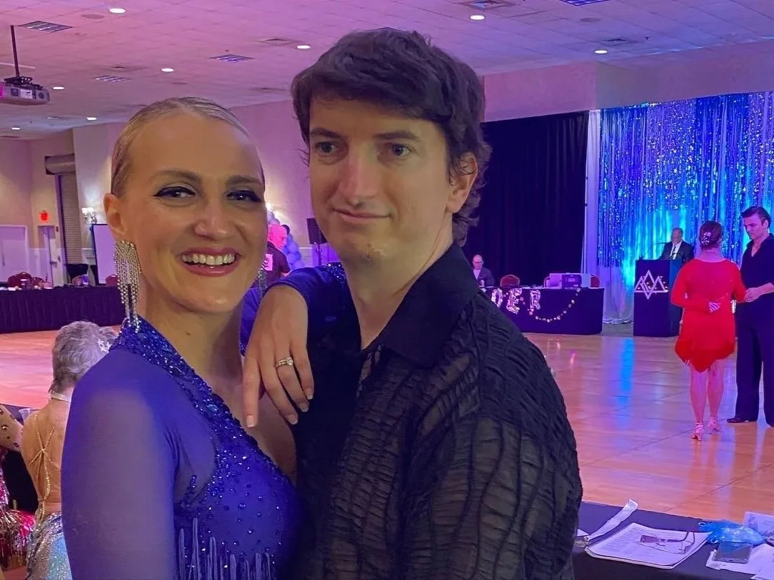 A couple dancing at a ballroom event, with people seated at tables in the background and a stage with a blue curtain and a band.