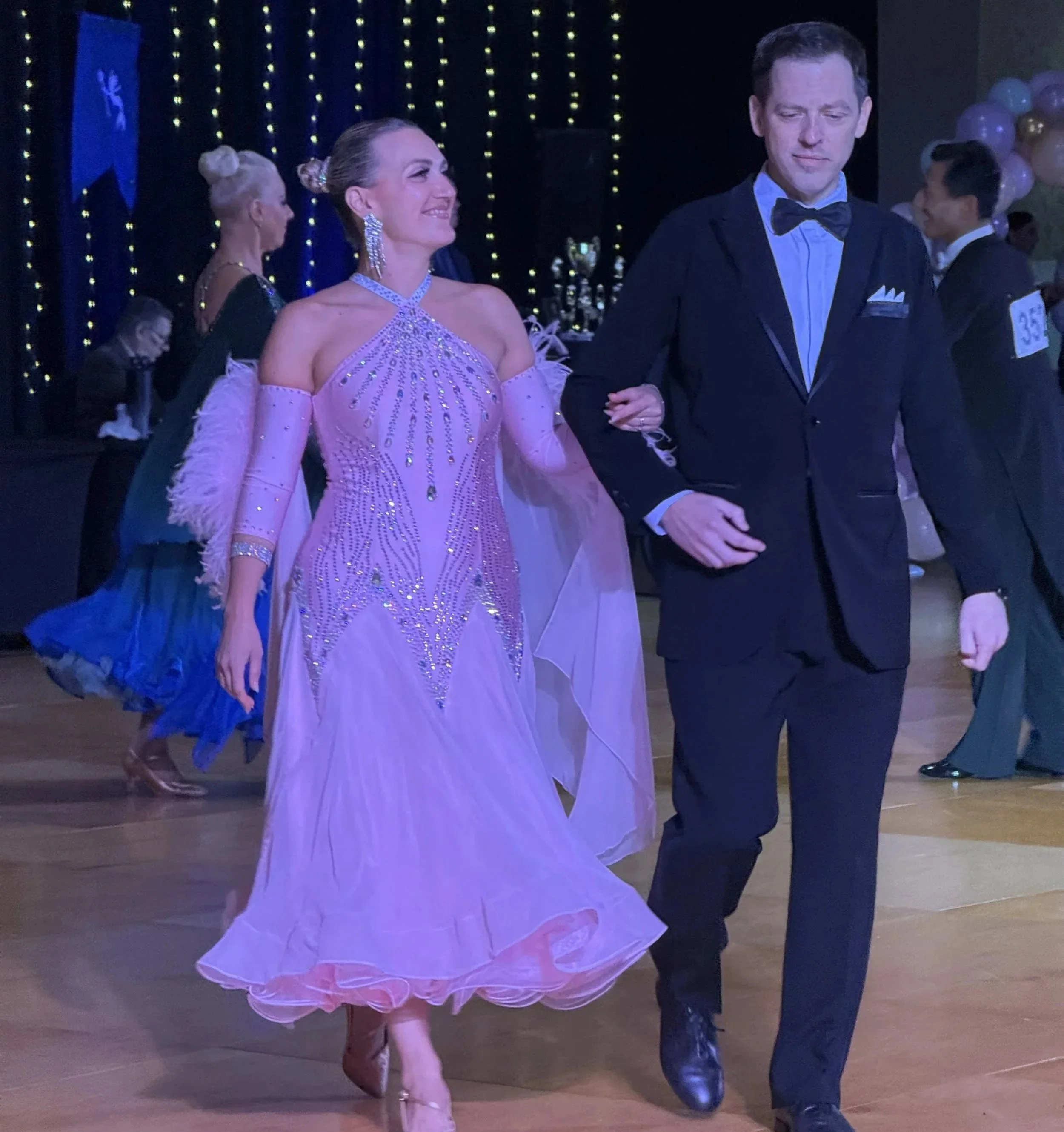 A woman in a fancy, lavender ballroom gown dancing with a man in a black tuxedo at a ballroom event.