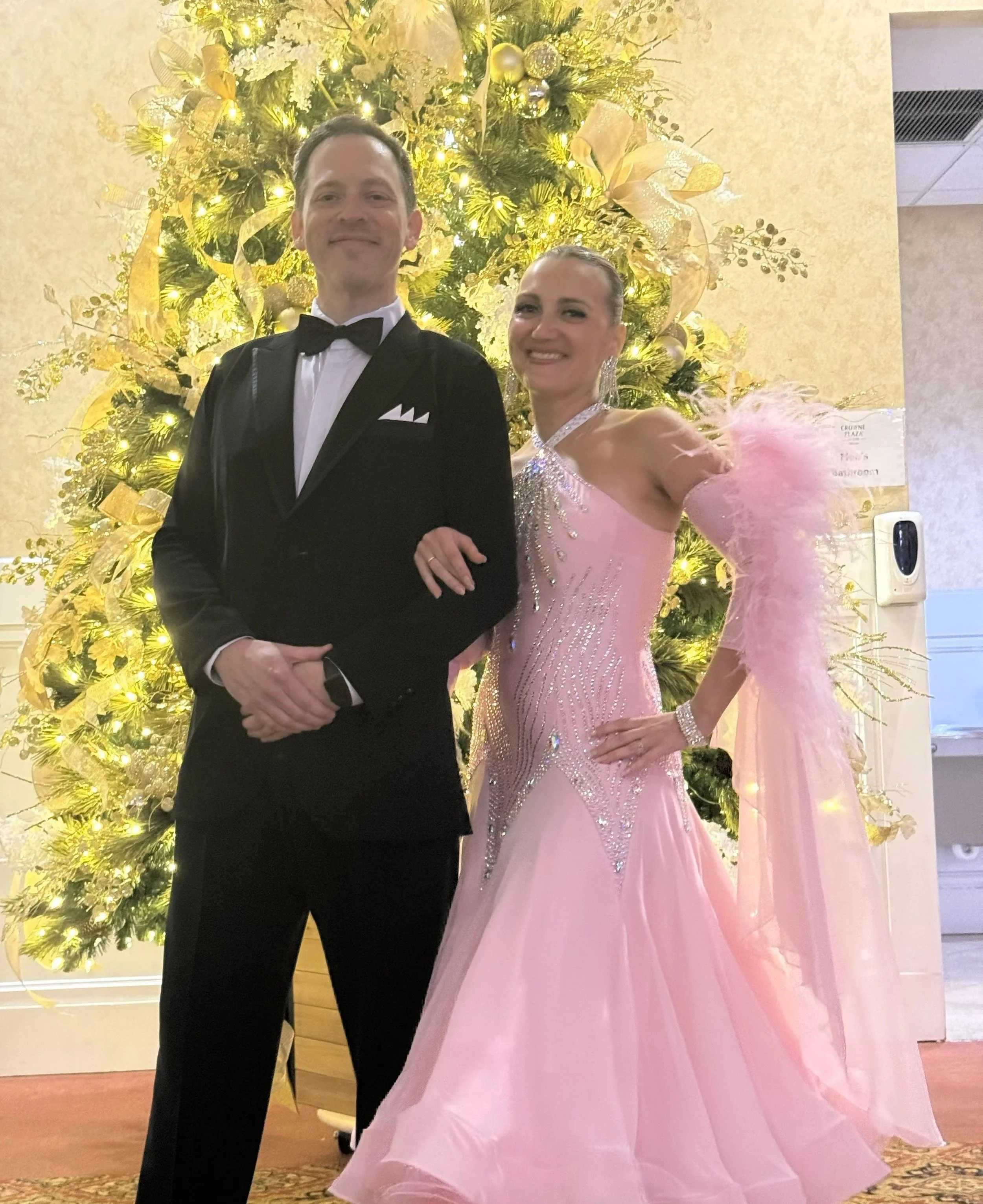 Man in black tuxedo and woman in pink ball gown with feathered sleeve standing in front of a decorated Christmas tree.