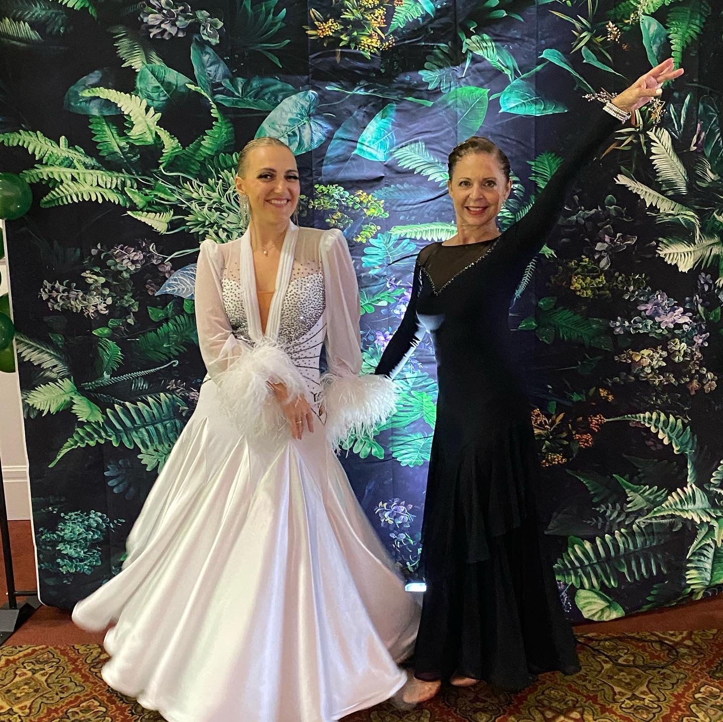 Two women dressed in elegant evening gowns, posing in front of a tropical-themed backdrop with large green leaves and flowers. The woman on the left wears a white gown with feathered cuffs, and the woman on the right wears a black gown with long slee