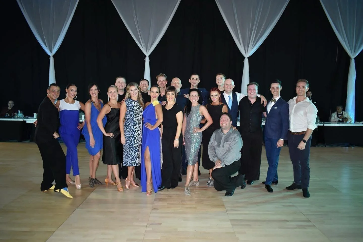 A group of 16 people in formal attire posing on a dance floor with black curtains and decorative drapes in the background.