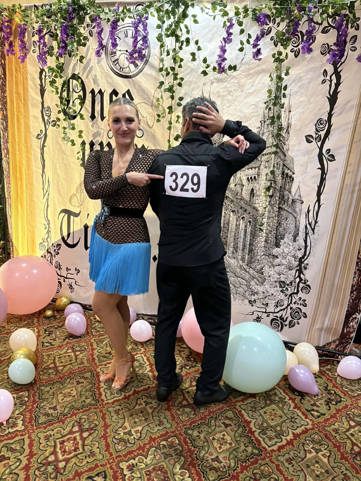 A woman in a black mesh top and blue fringe skirt dances with a man wearing a black shirt numbered 329 at a themed event with a castle backdrop, hanging purple flowers, and pastel balloons on the carpeted floor.