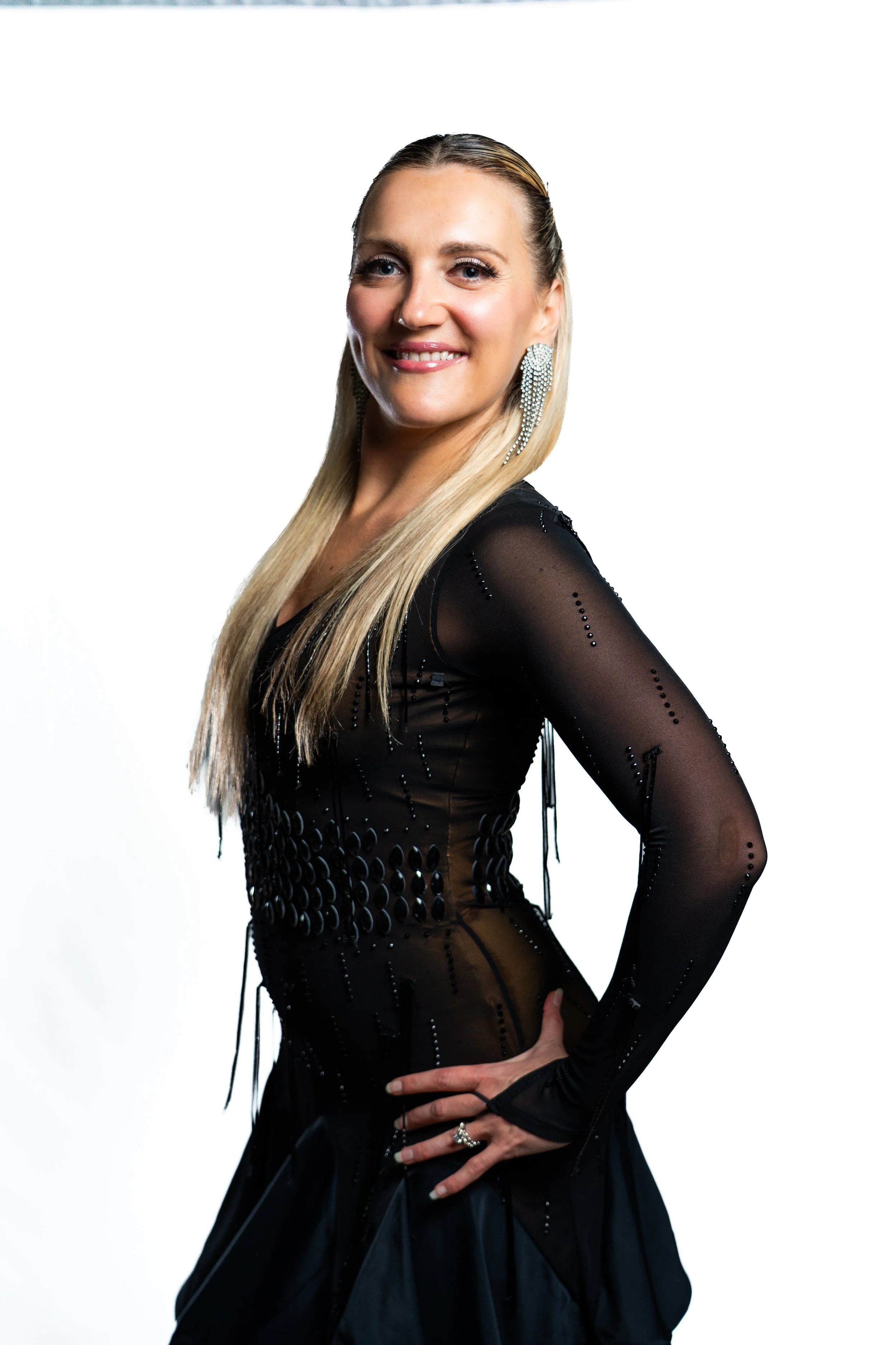 A woman with blonde hair, smiling, wearing a black dress with sheer long sleeves and silver earrings, posing against a white background.