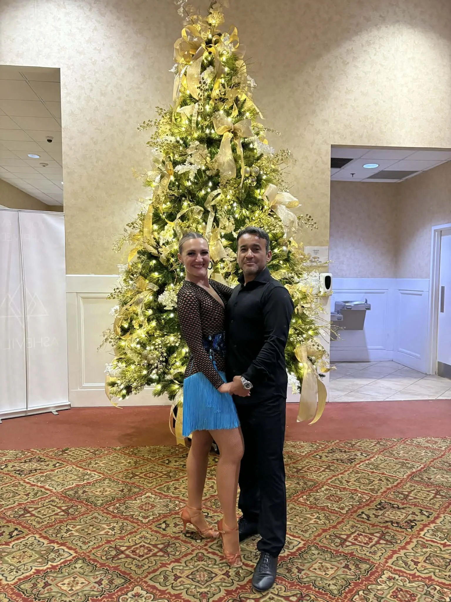 A couple dancing in front of a decorated Christmas tree with gold ribbons and lights.