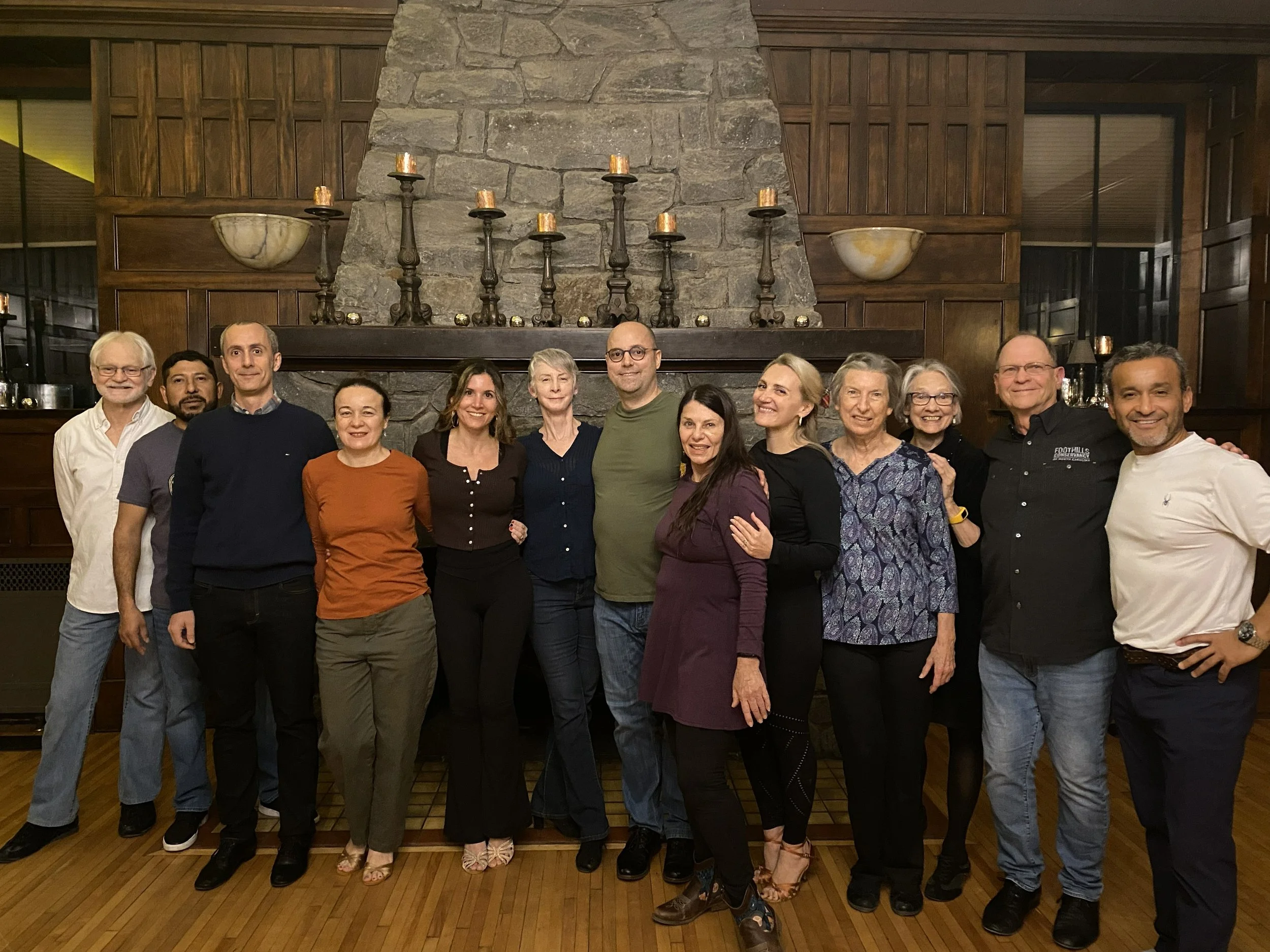 Group of 14 people standing together in a warmly lit room with a stone fireplace and candles.