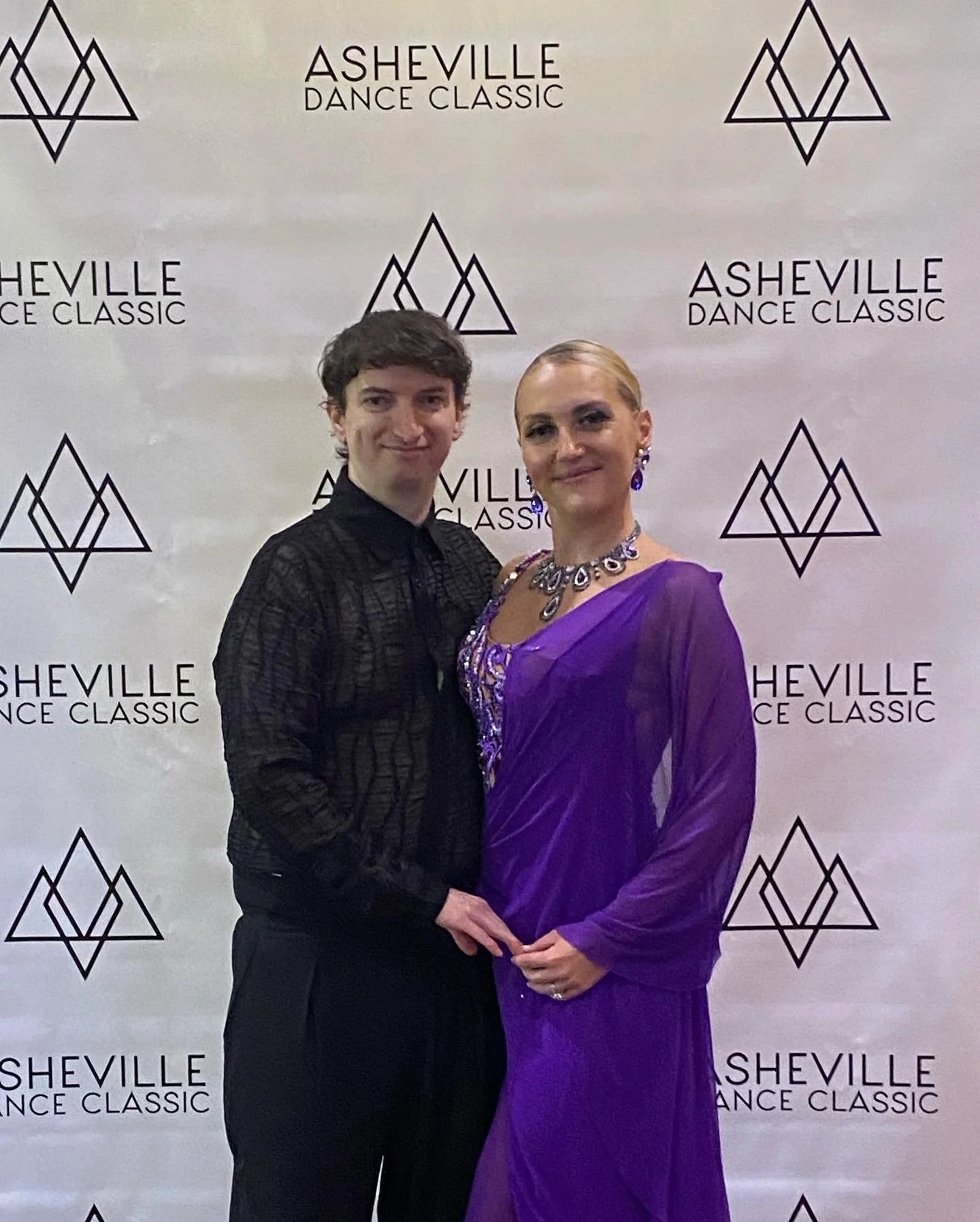 A man and a woman standing together at the Asheville Dance Classic event, posing in front of a backdrop with the event's logo.