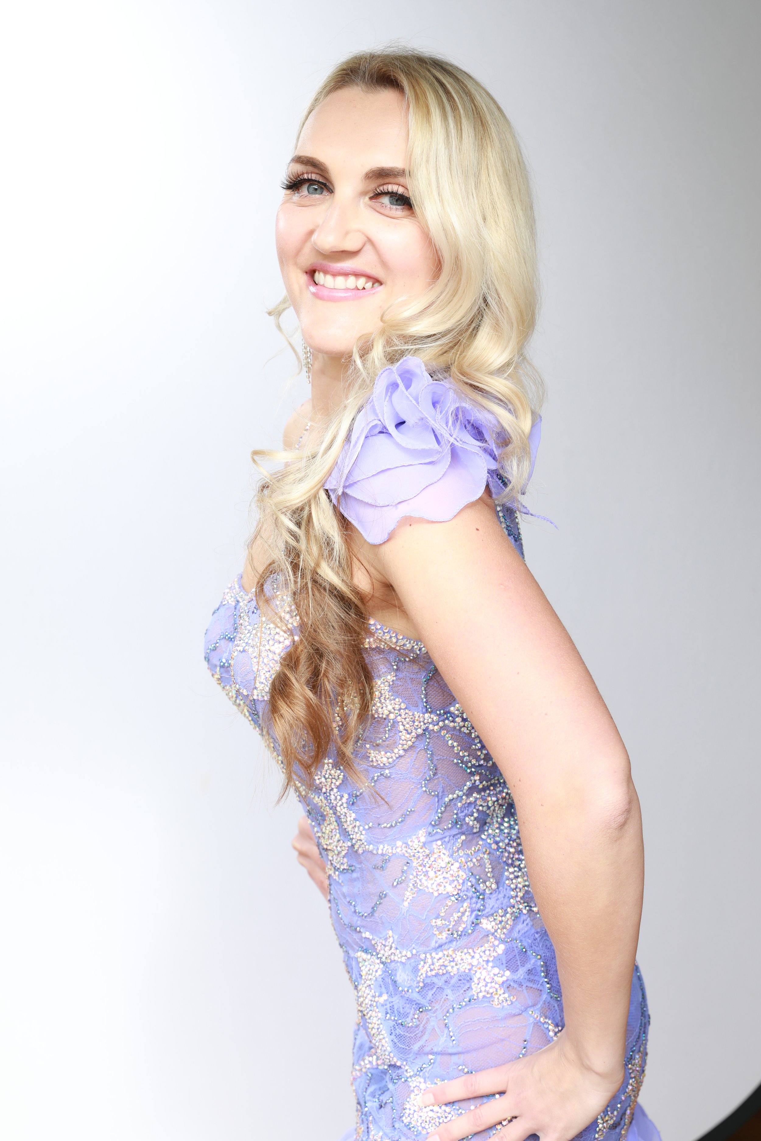 A smiling woman with long blonde curly hair wearing a lavender dress with ruffled sleeve and bead embellishments, standing against a plain background.