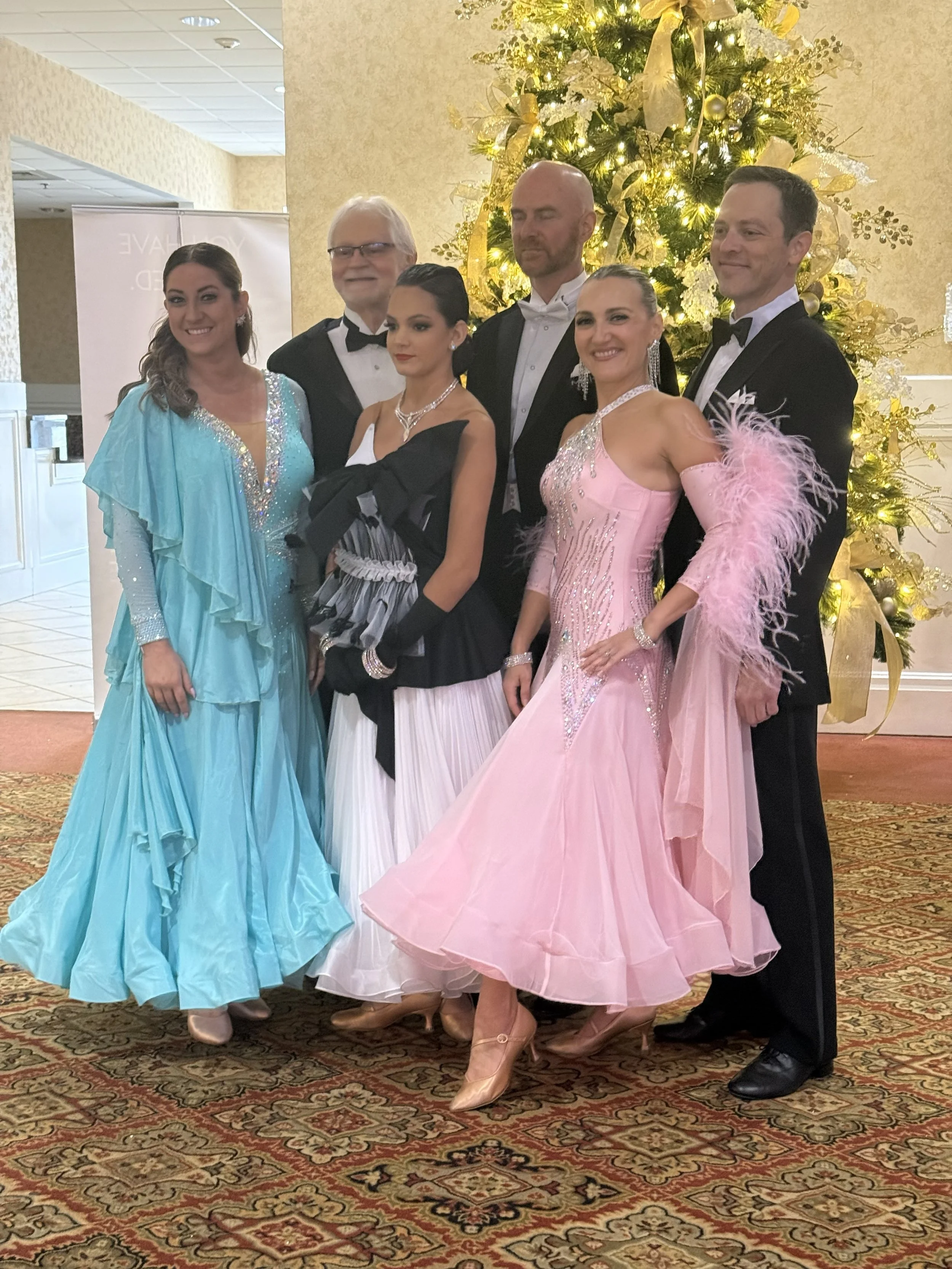 Group of six people dressed in formal attire, standing in front of a decorated Christmas tree with gold ribbons and lights, in an indoor setting with a patterned carpet.