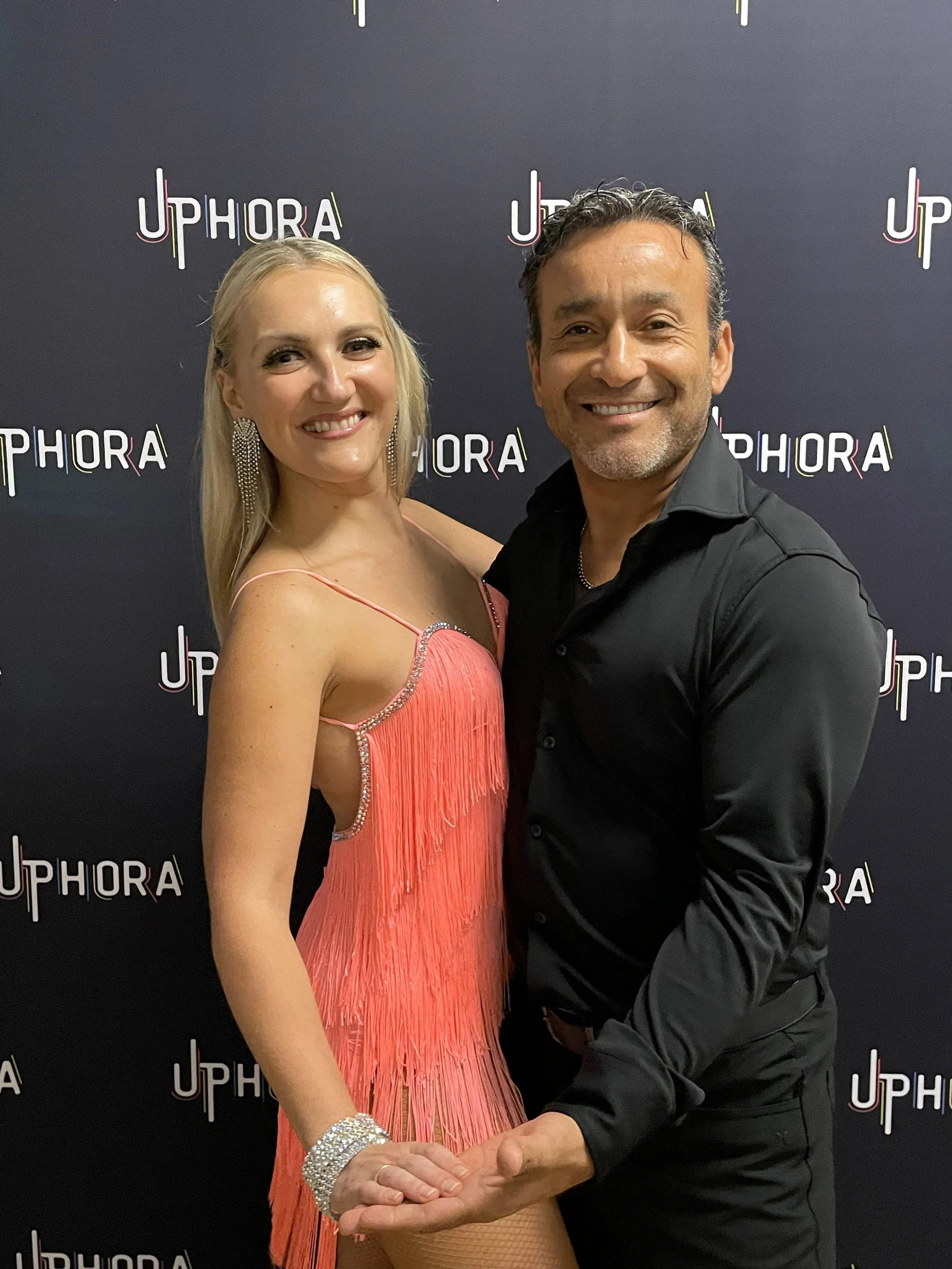 A man and woman pose together at a Sephora event, smiling, with the Sephora logo on the black backdrop behind them. The woman is wearing a pink dress with fringes and statement jewelry, and the man is dressed in a black shirt.
