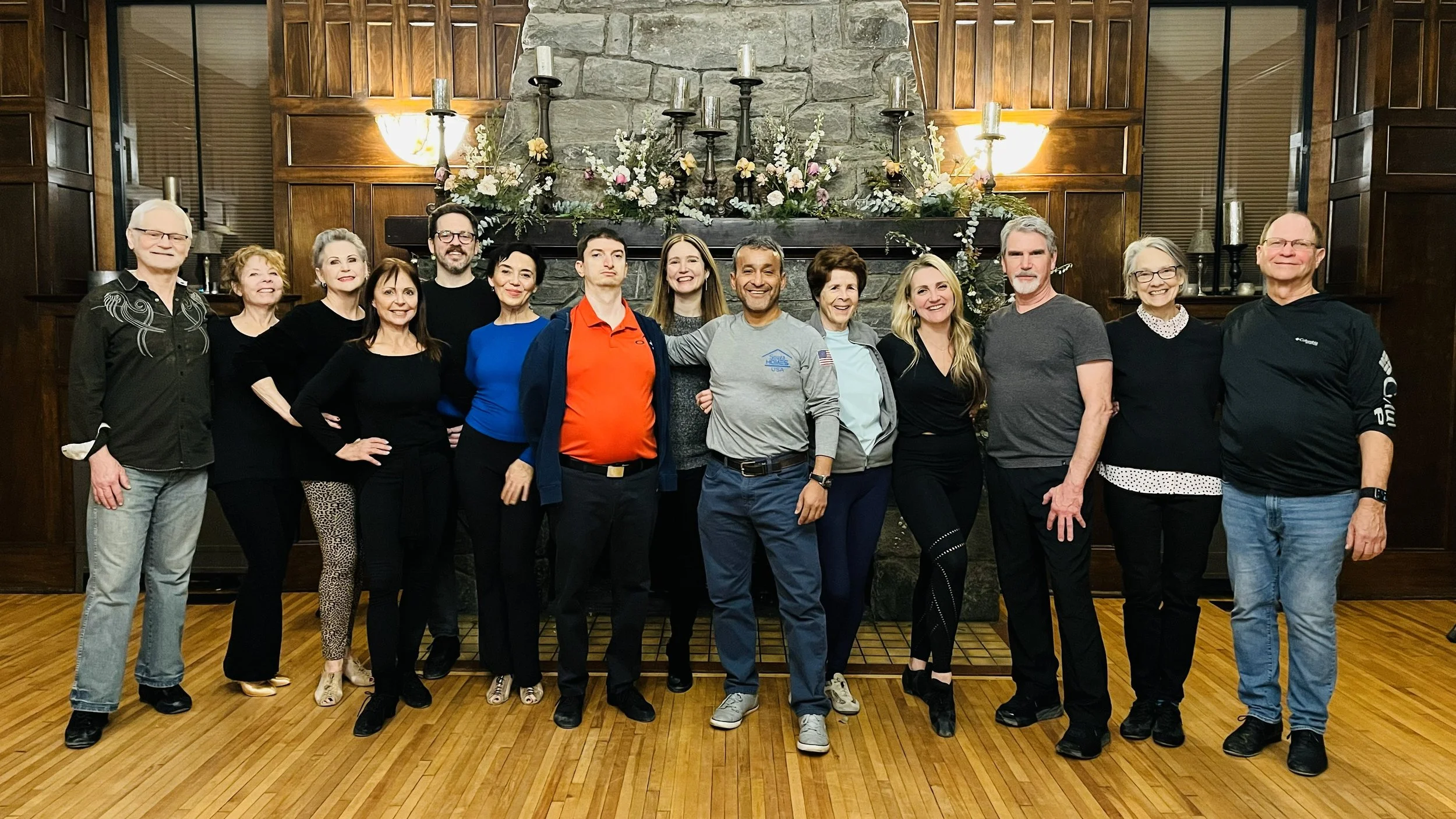 A group of fifteen people posing near a stone fireplace with floral decoration in a wood-paneled room, smiling for the photo.