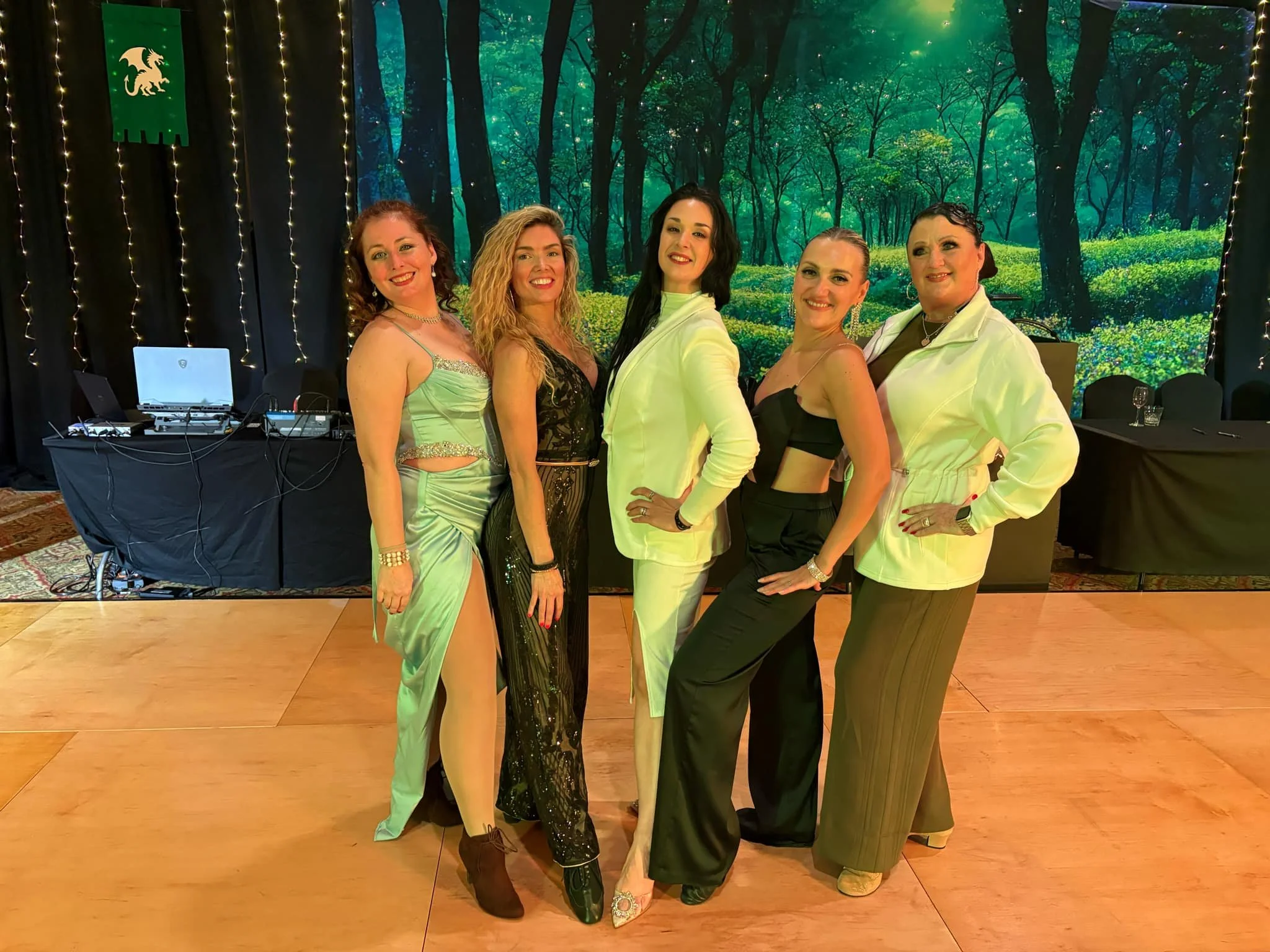 Five women standing together on a dance floor, dressed in elegant and stylish outfits, with a forest-themed backdrop and fairy lights behind them.