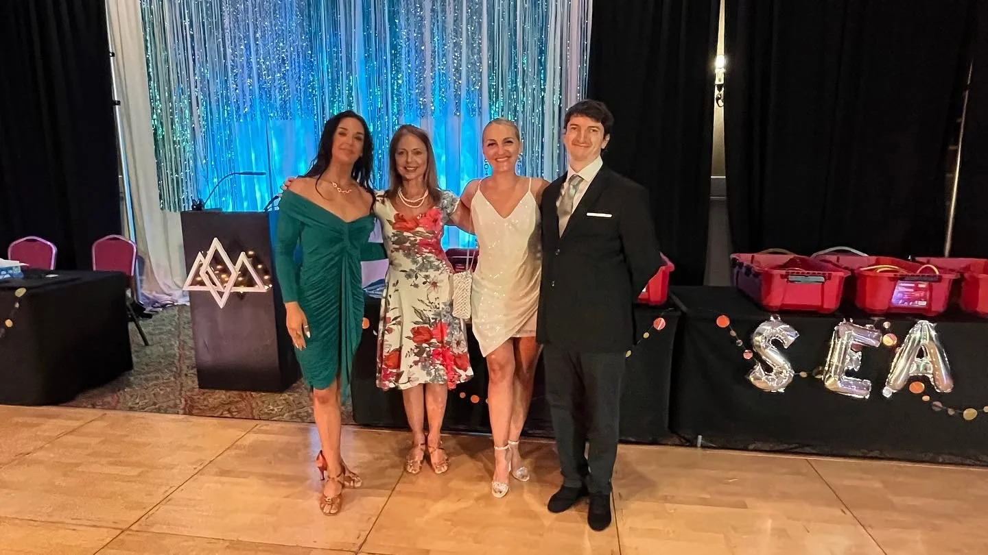 Group of four people in formal attire standing on a dance floor at an event, with decorative backdrop and a table with red gift bags and "SEA" spelled out in balloons.