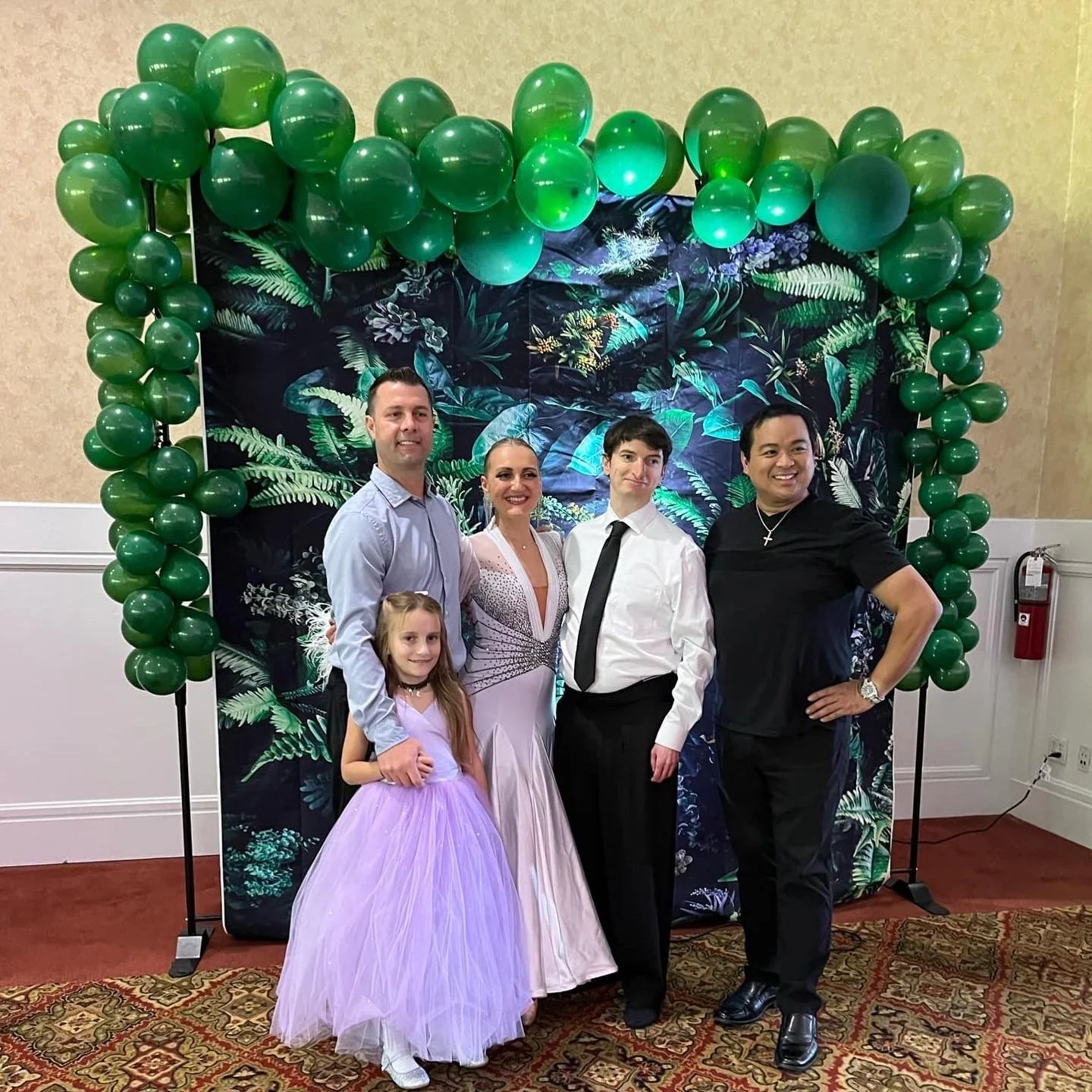 Group of five people standing in front of a tropical-themed backdrop with green balloon arch at a celebration or event. Two men, one woman, one girl in a pink dress, and one woman in a white dress.