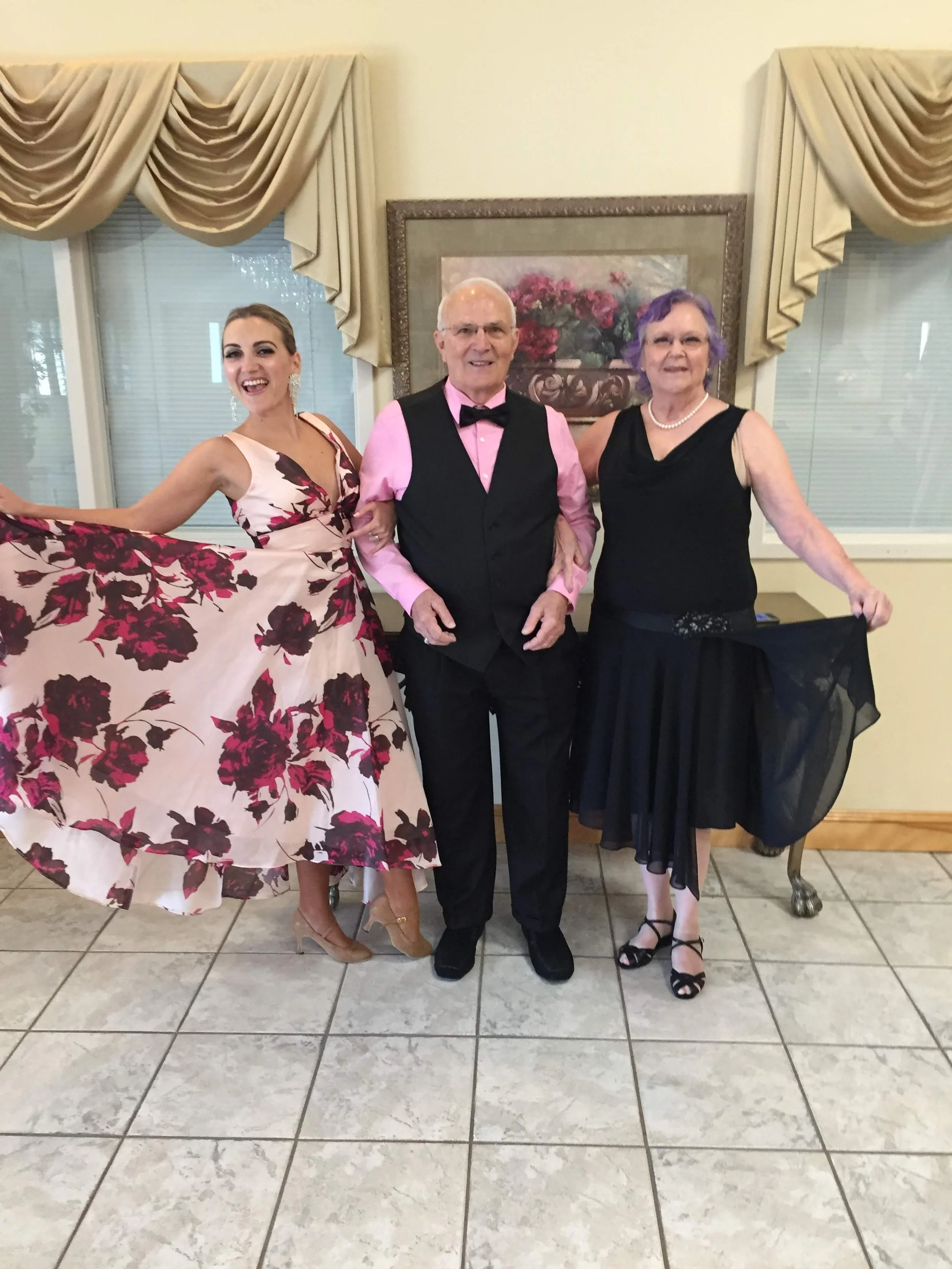 Three people in formal attire are posing together indoors. The woman on the left is wearing a beige dress with a large floral pattern and beige heels. The man in the middle is wearing a pink shirt, black vest, black pants, and a pink bow tie. The wom