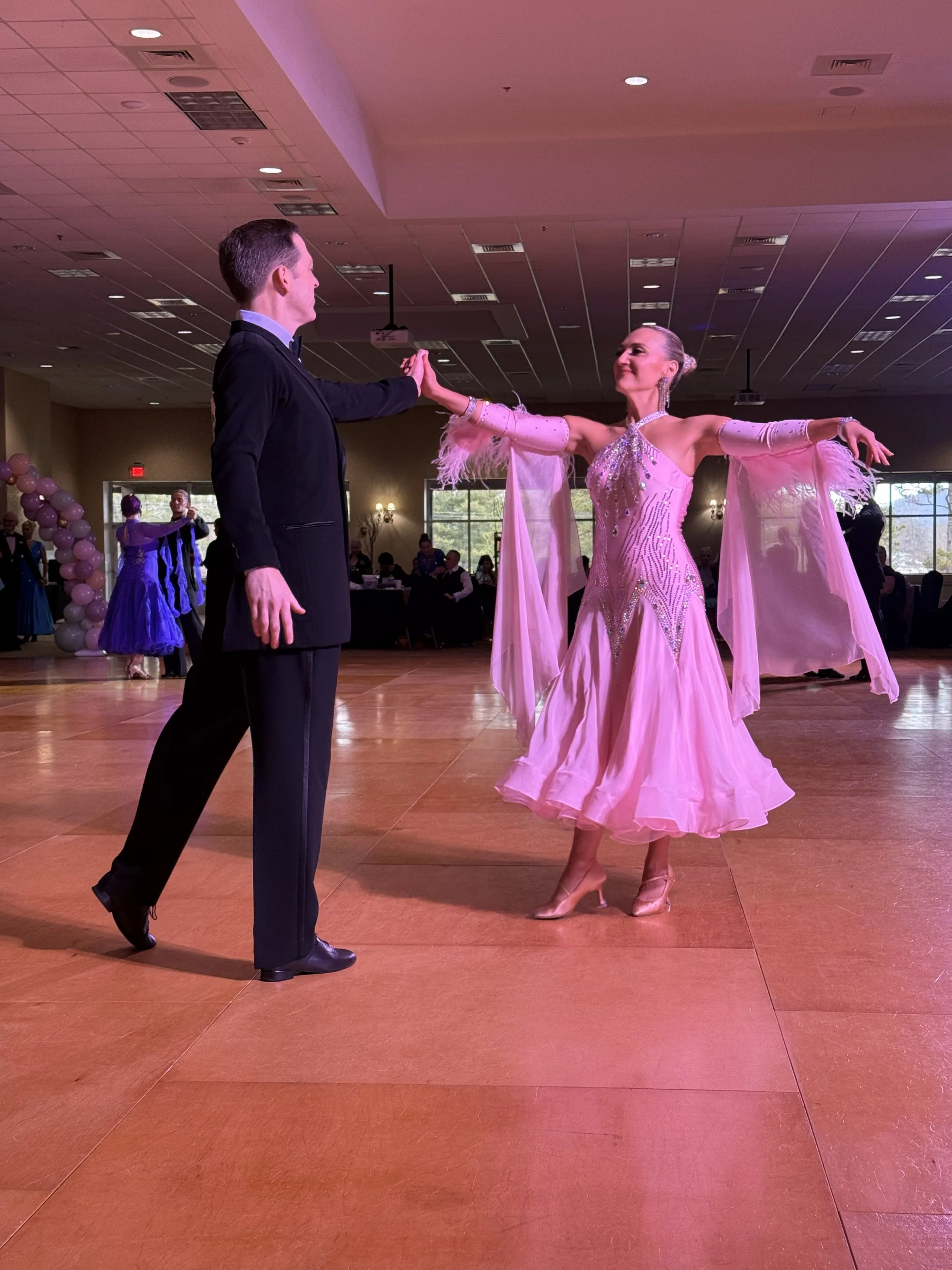 A couple dancing ballroom dance in a large decorated venue with pink lighting and guests sitting at tables in the background.