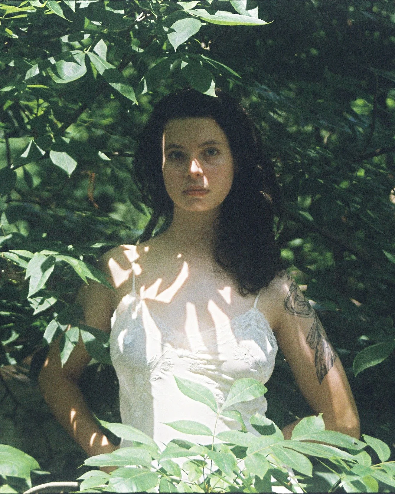 A woman with dark curly hair and tattoos on her arm, standing among green leaves. She has a neutral expression, wears a white lace dress, and is partially illuminated by sunlight filtering through the leaves.