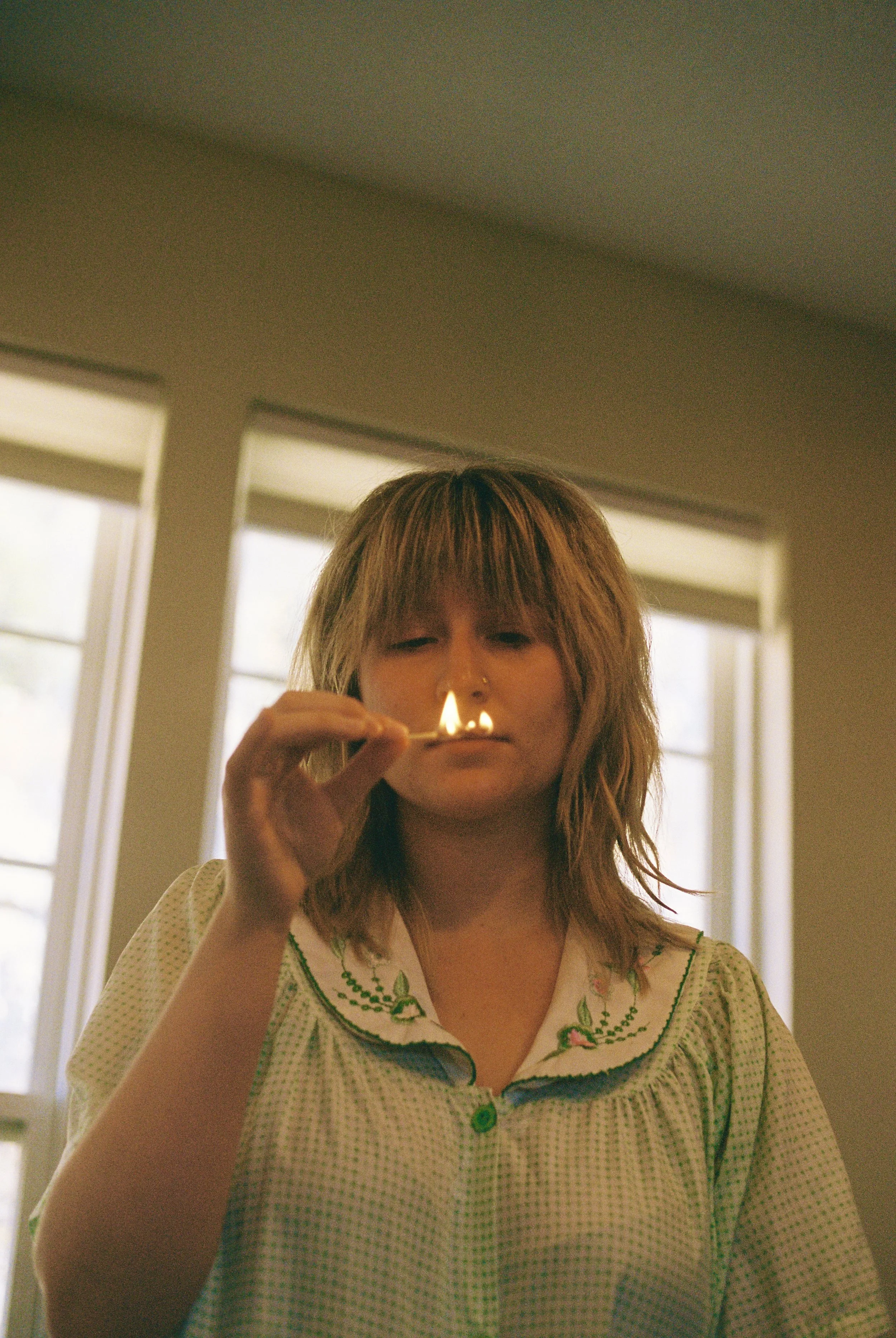 A woman with shoulder-length brown hair, wearing a light-colored, embroidered nightgown, is looking down at a lit match she is holding in front of her face.