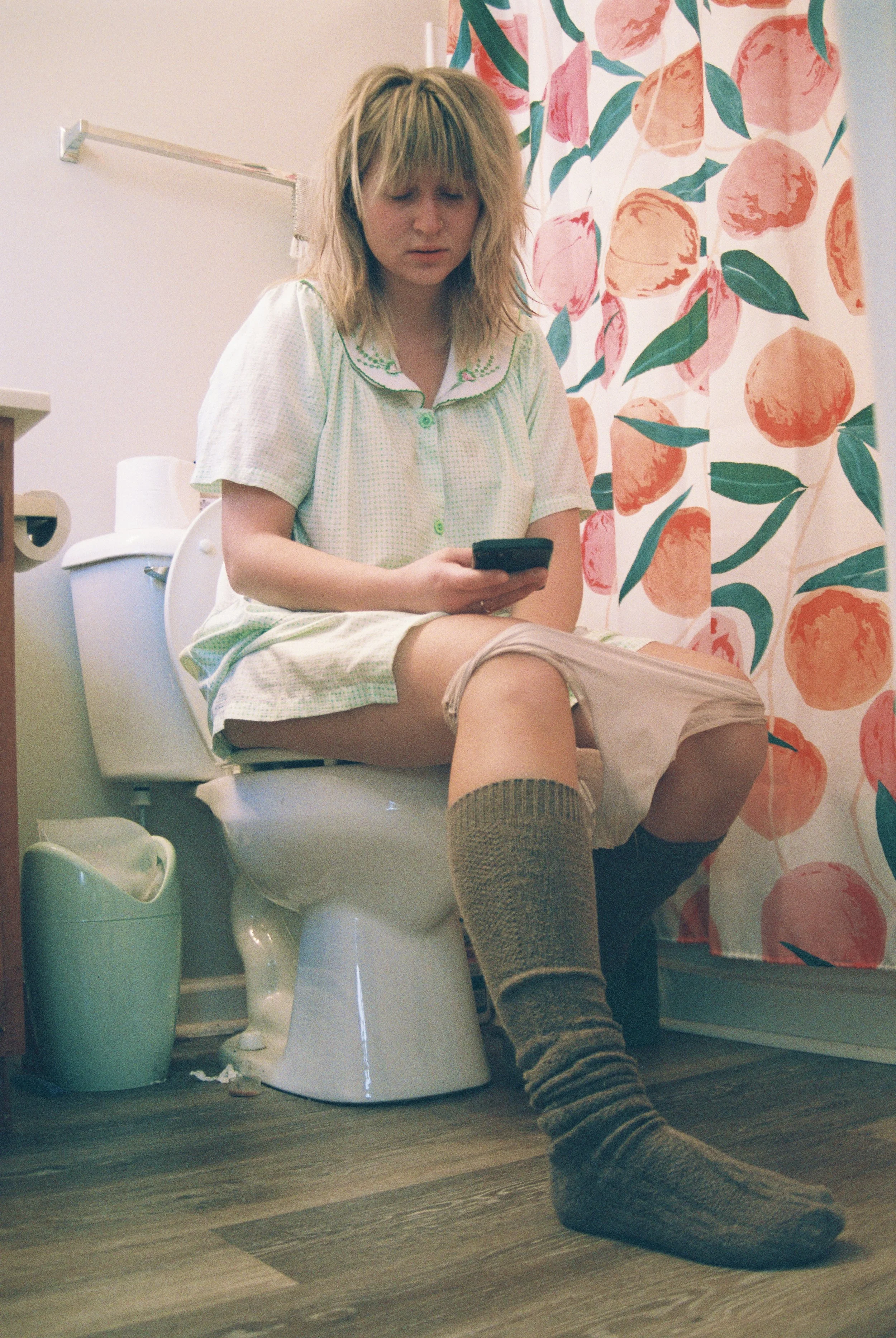 A woman with blond hair is sitting on a toilet in a bathroom, looking at her phone. She is wearing a loose nightgown and long socks. The bathroom has a floral shower curtain with pink and orange flowers and green leaves, and a trash bin beside the to