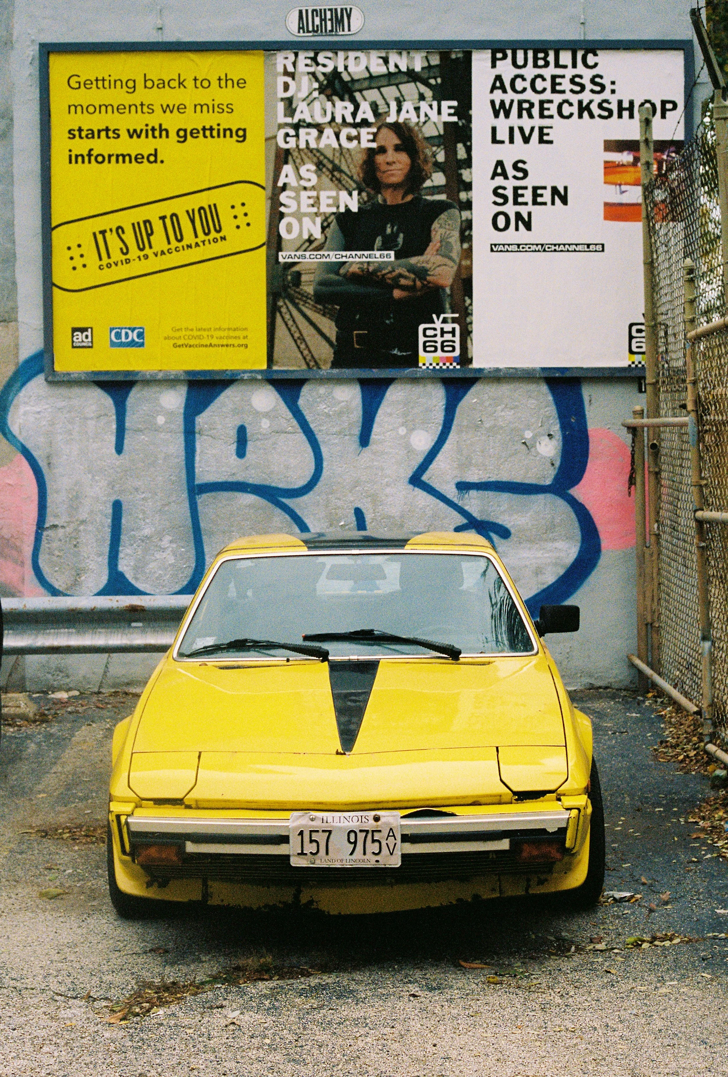 A yellow vintage car with a black stripe on the hood, parked in front of a graffiti wall and a large billboard. The billboard contains various advertisements and images, including a woman and public health messages.