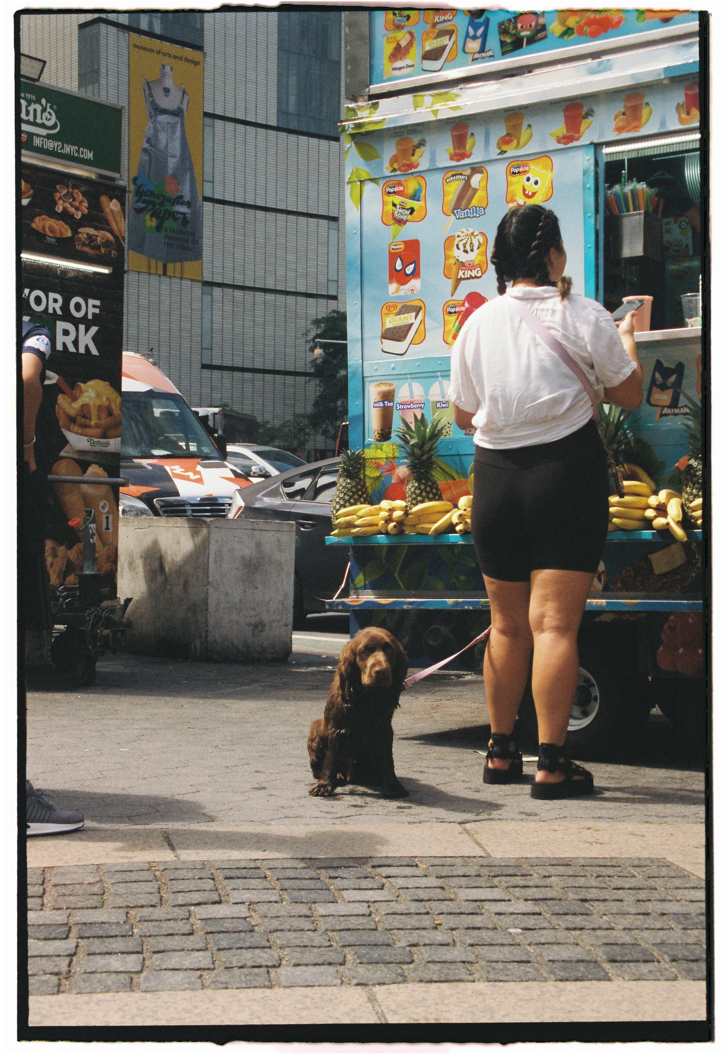 A woman with dark hair in braids, wearing a white shirt and black shorts, standing at a juice and smoothie stand with bananas and pineapples, holding a smartphone. A brown dog on a pink leash sits on the sidewalk nearby. Behind the woman, there's a c