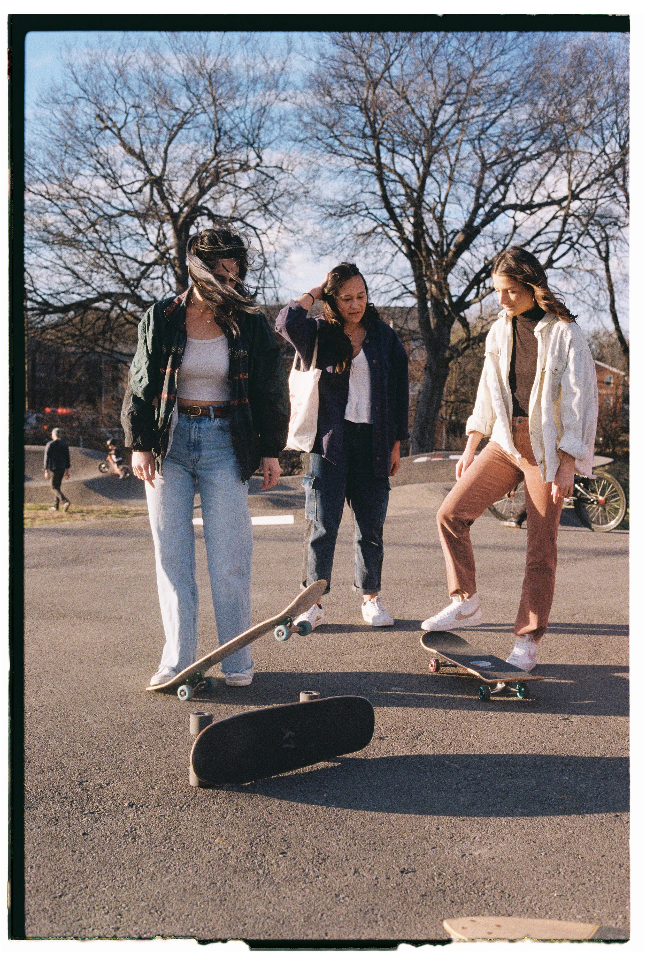 Four young women at a skatepark, with two skateboards on the ground, during daytime with bare trees in the background.