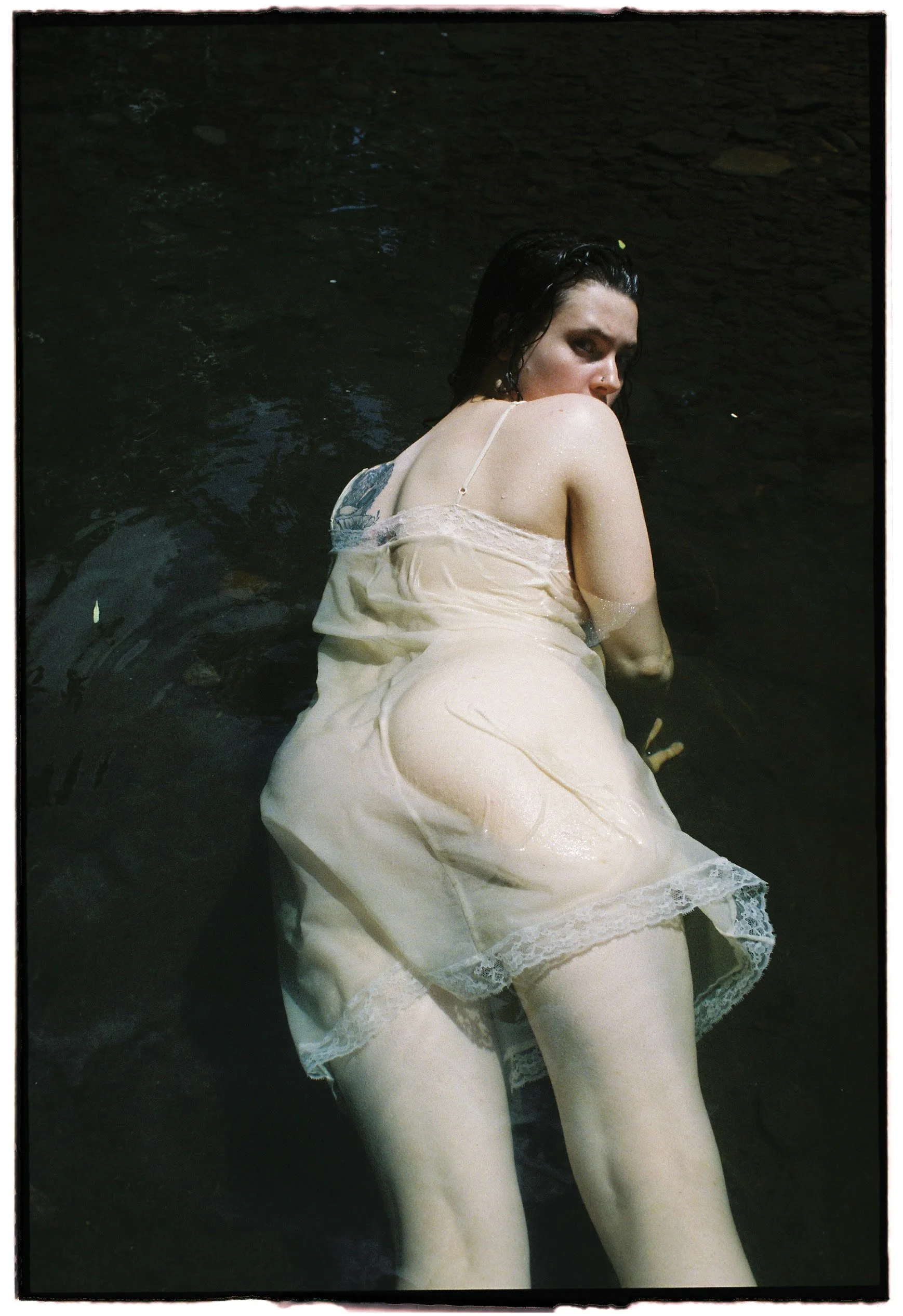 A woman lying in a river, wearing a light-colored, wet nightgown with lace trim, gazing over her shoulder at the camera.