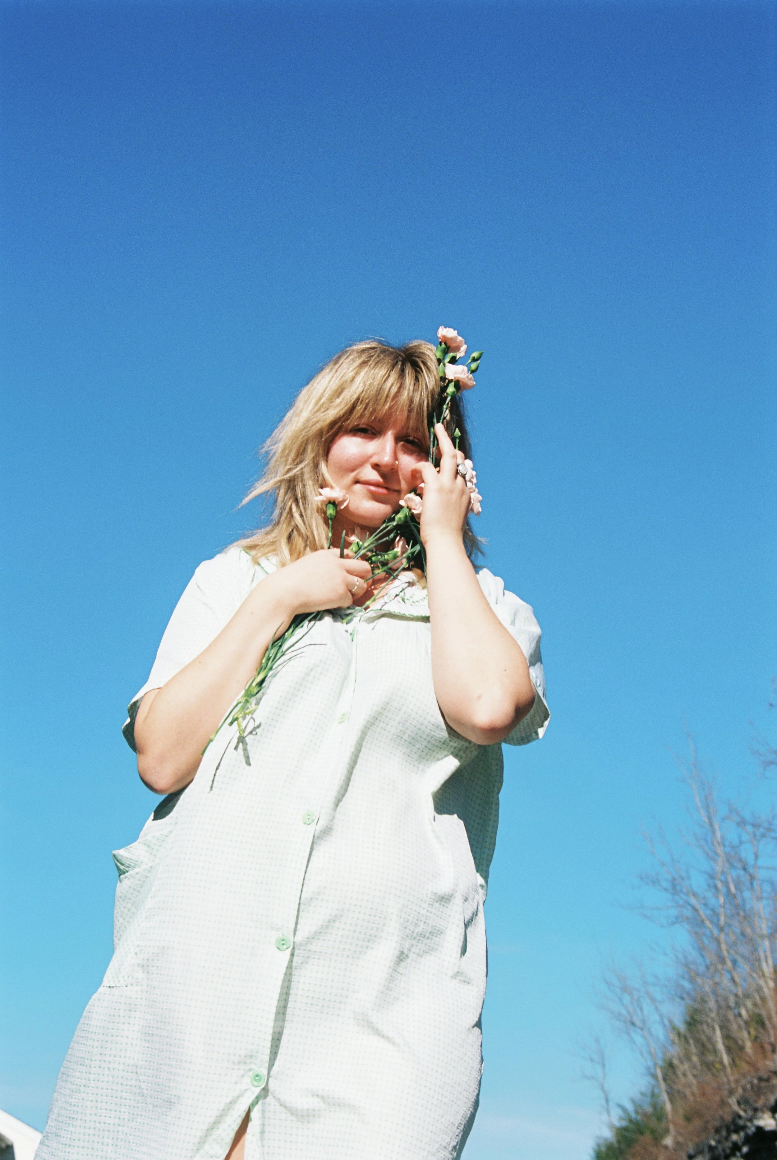 A woman with blonde hair holding pink flowers against a clear blue sky.