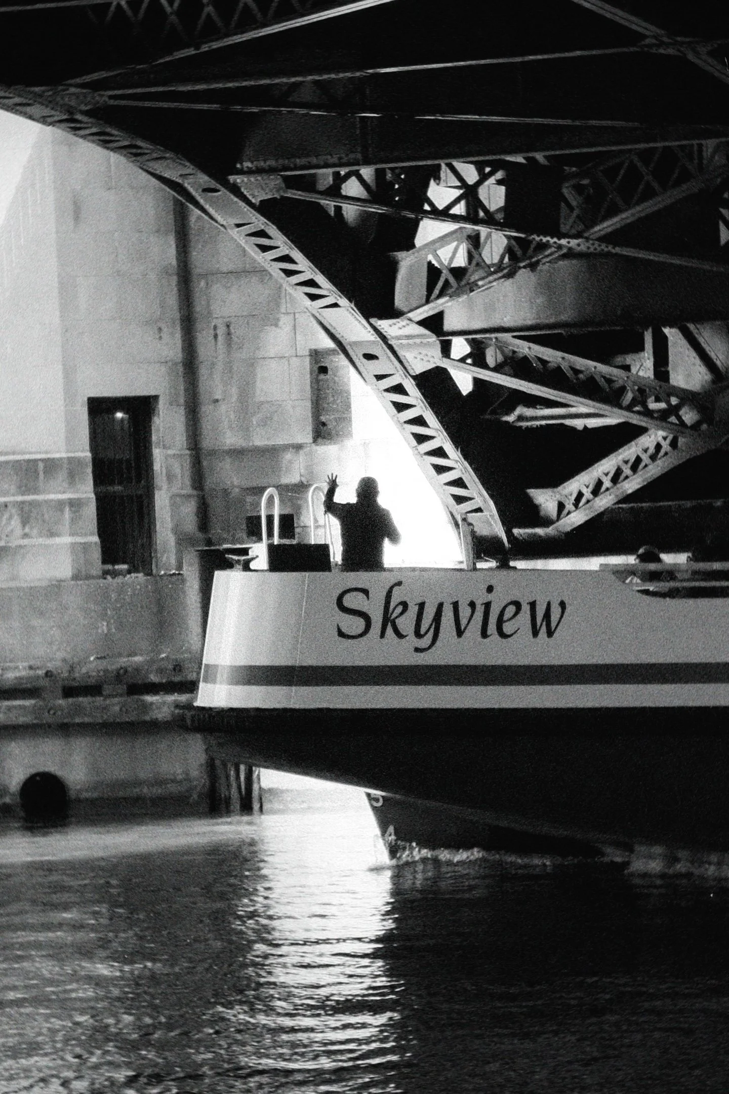 A person is standing on the edge of a boat named Skyview, waving, with a large steel bridge structure in the background, under low lighting conditions.