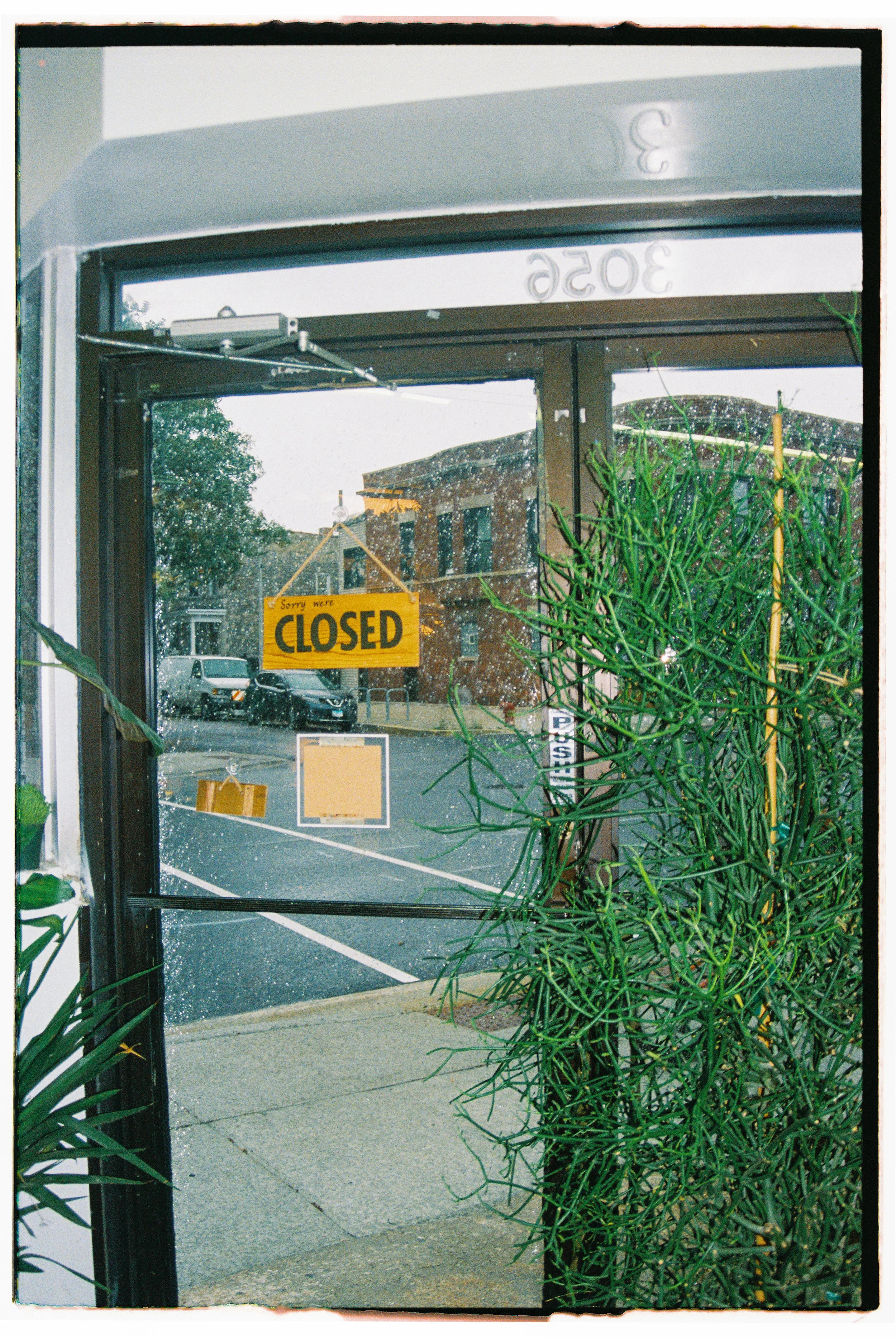 Rain-speckled glass door with a yellow 'Sorry we' closed' sign hanging, next to green leafy plants, and an outdoor parking lot visible through the glass.