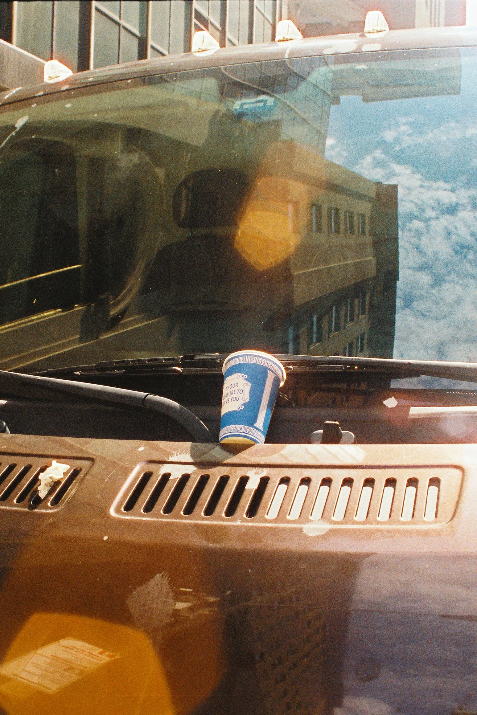 View from inside a vehicle looking through the windshield at a city scene with a tall building, a blue disposable cup on the dashboard, and satellite lights on the roof of the vehicle.