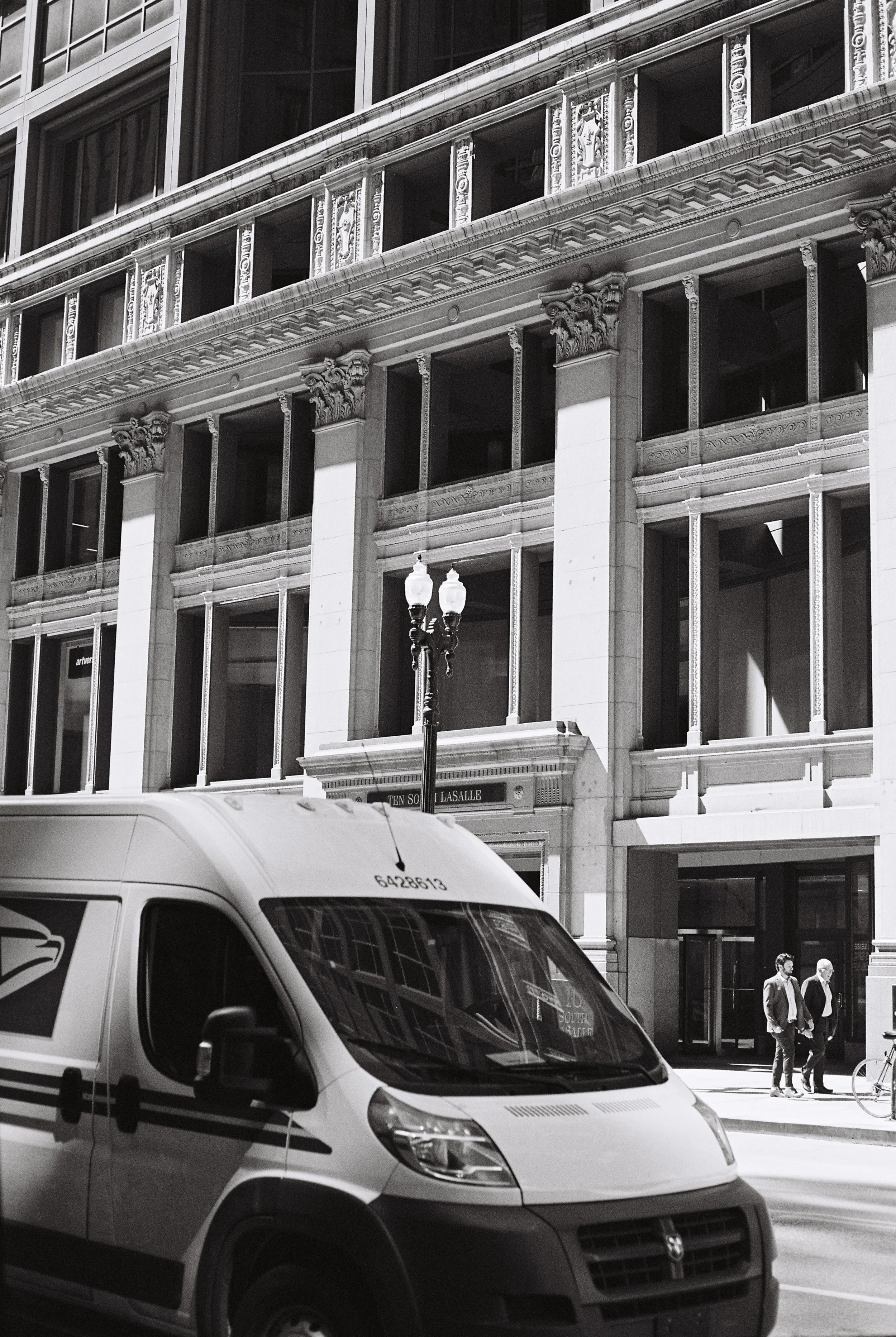 Street scene with a white postal delivery van parked on the side of the road, a historic building with ornate architectural details, and two people walking on the sidewalk at night.