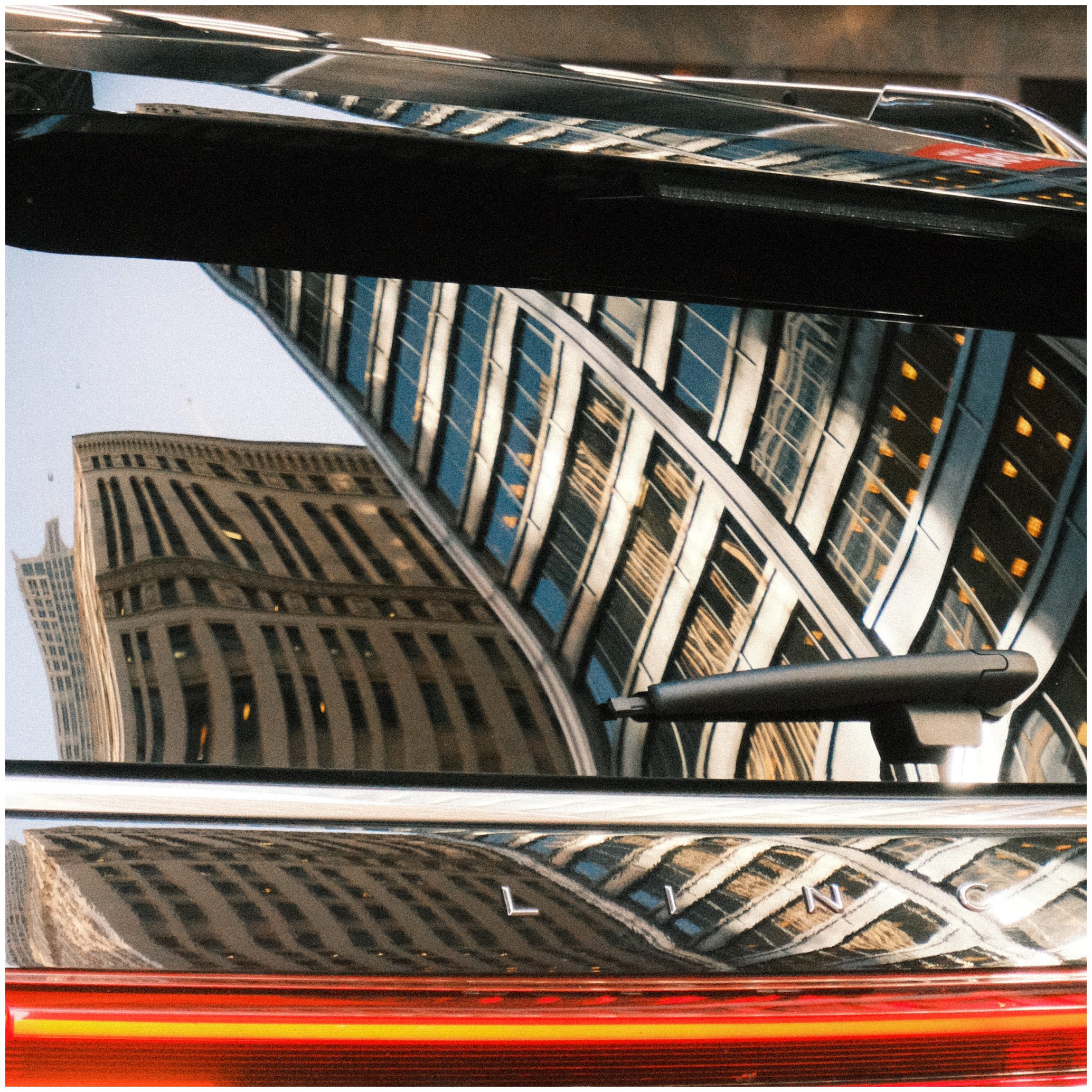 Reflection of tall office buildings on a shiny black car's trunk with Lincoln logo, with the photo taken at an upward angle.