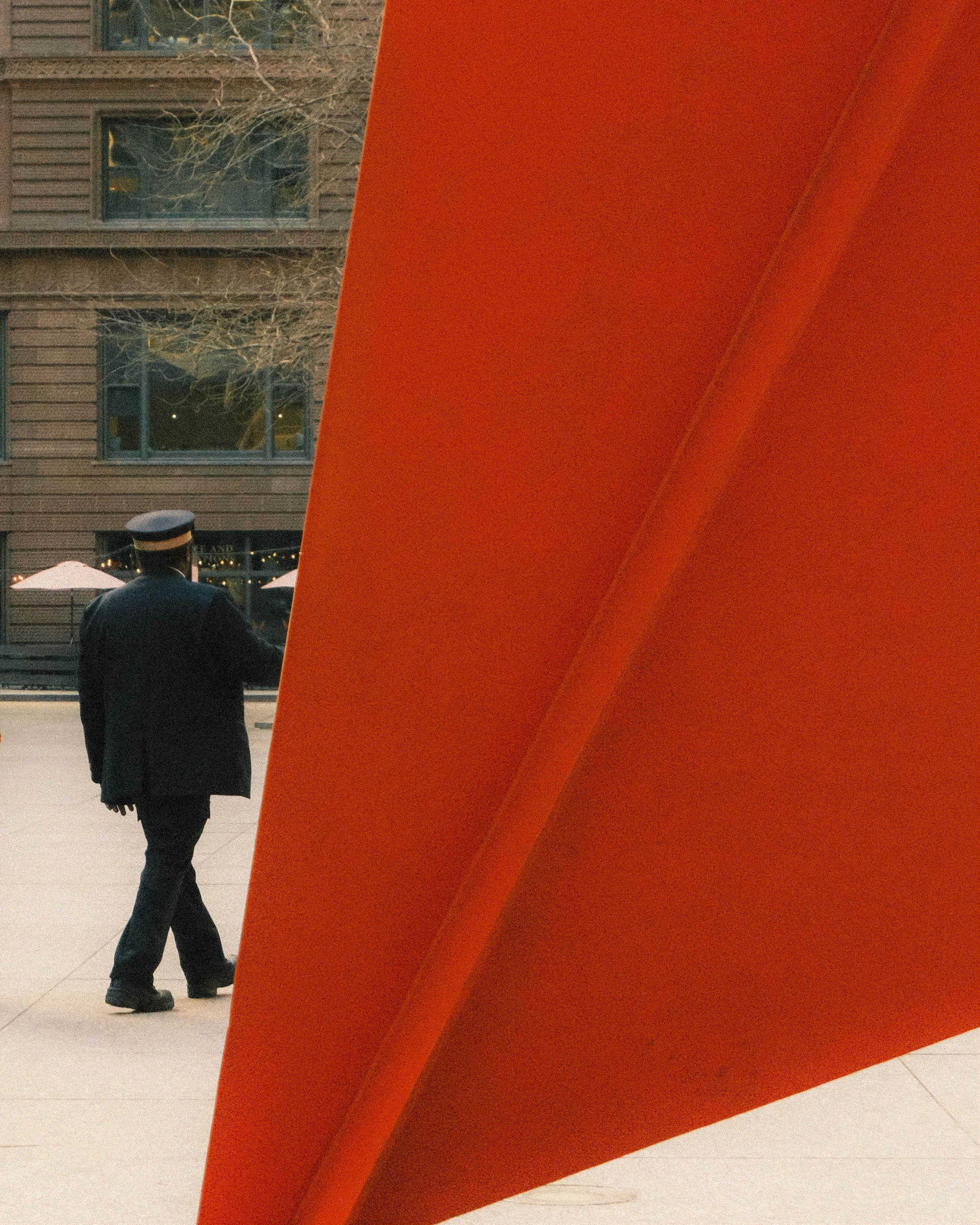 A man in a black jacket and hat is walking past a large red flamingo sculpture by Alexander Calder in Chicago. There are buildings and outdoor umbrellas in the background.