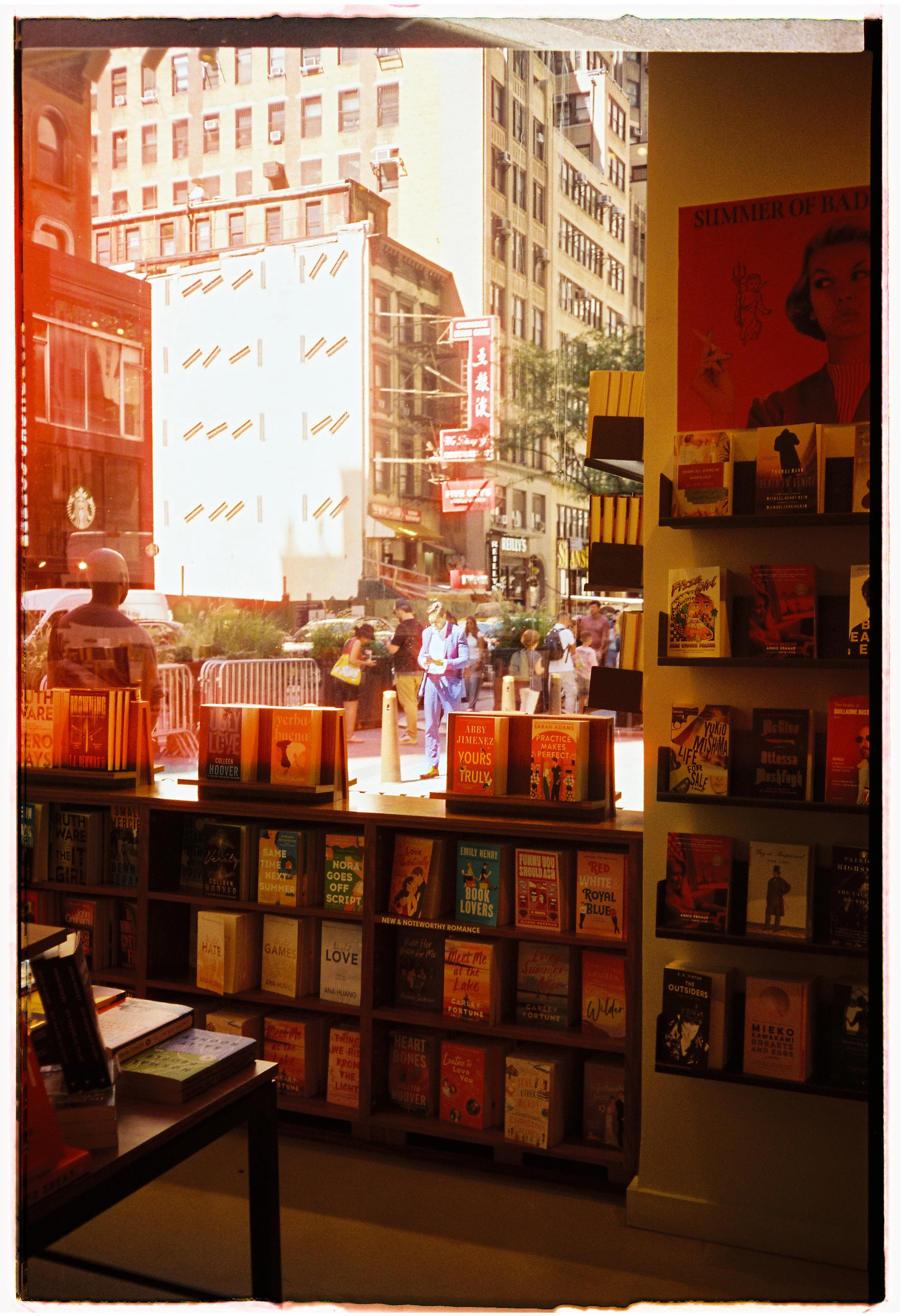 View of a bookstore shelf with books inside, looking out onto a busy city street with pedestrians and tall buildings.