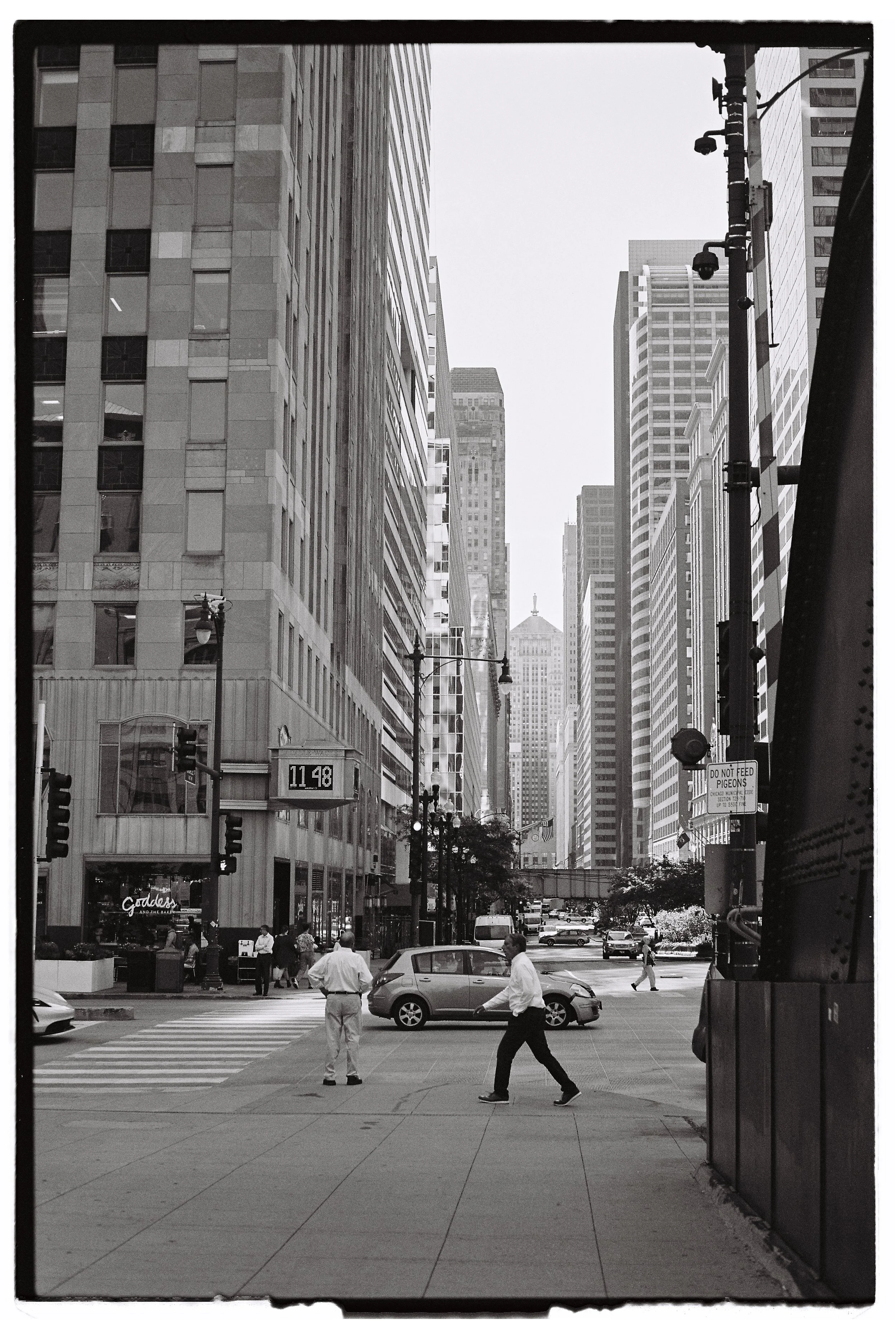Black and white photo of a busy city street with tall skyscrapers, pedestrians crossing, and cars on the road, including a traffic light and a sign that reads 'Do not feed pigeons'.