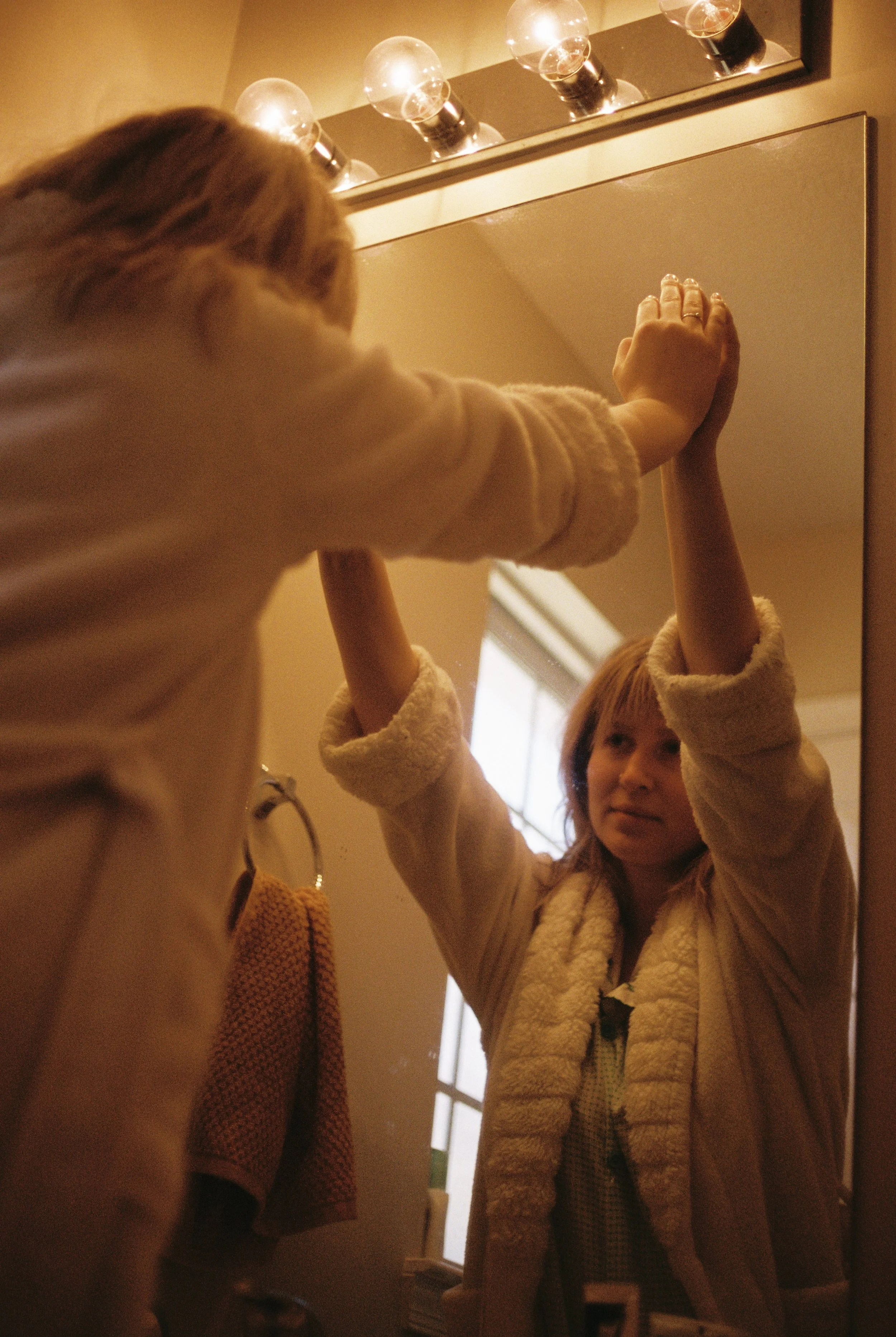 A woman looks at her reflection in a mirror as her child touches the mirror with both hands, reaching above her head. Both are dressed in cozy, light-colored sweaters, and there is a towel hanging on a hook next to the mirror.