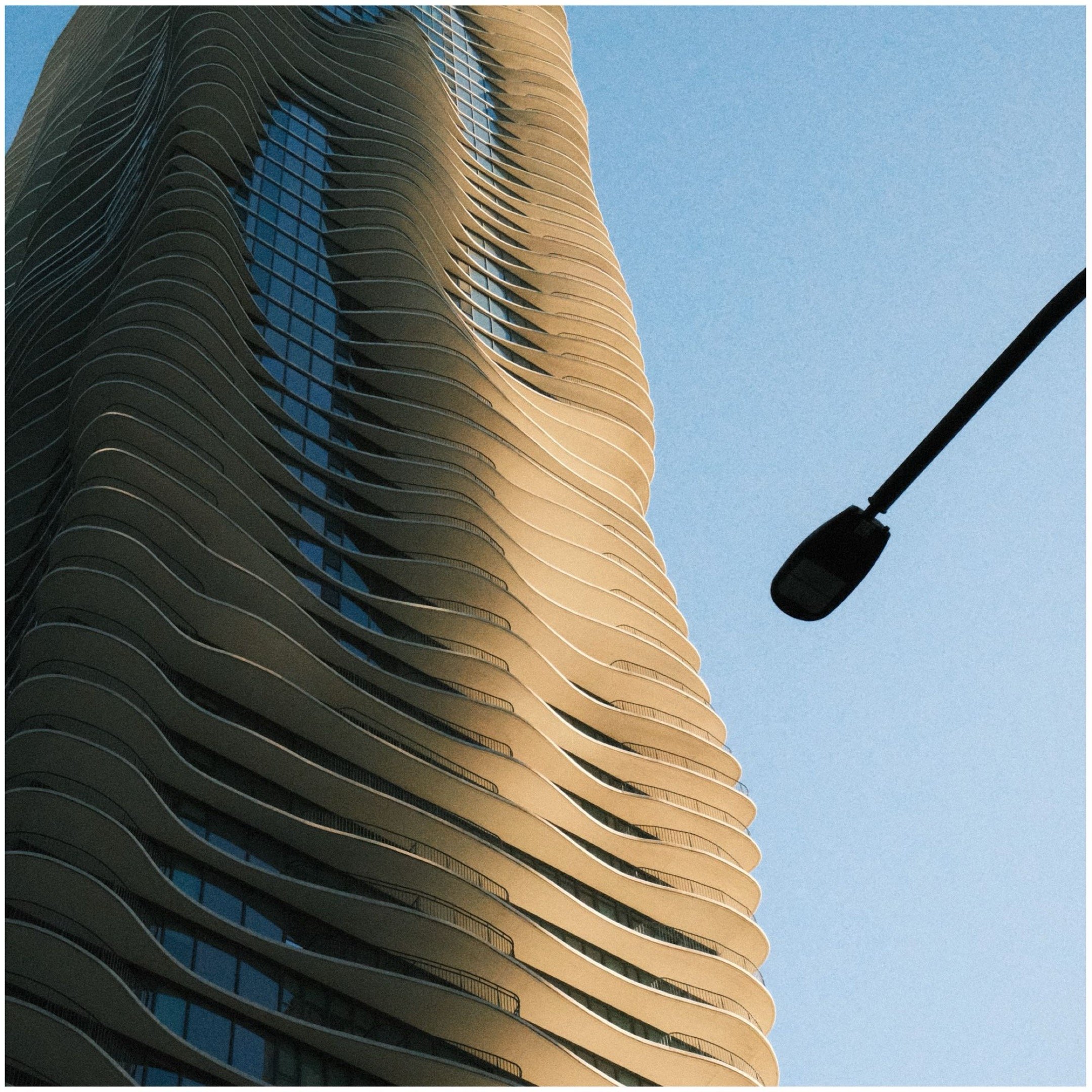 Low-angle view of a modern high-rise building with wavy balconies, and a streetlight against a clear blue sky.