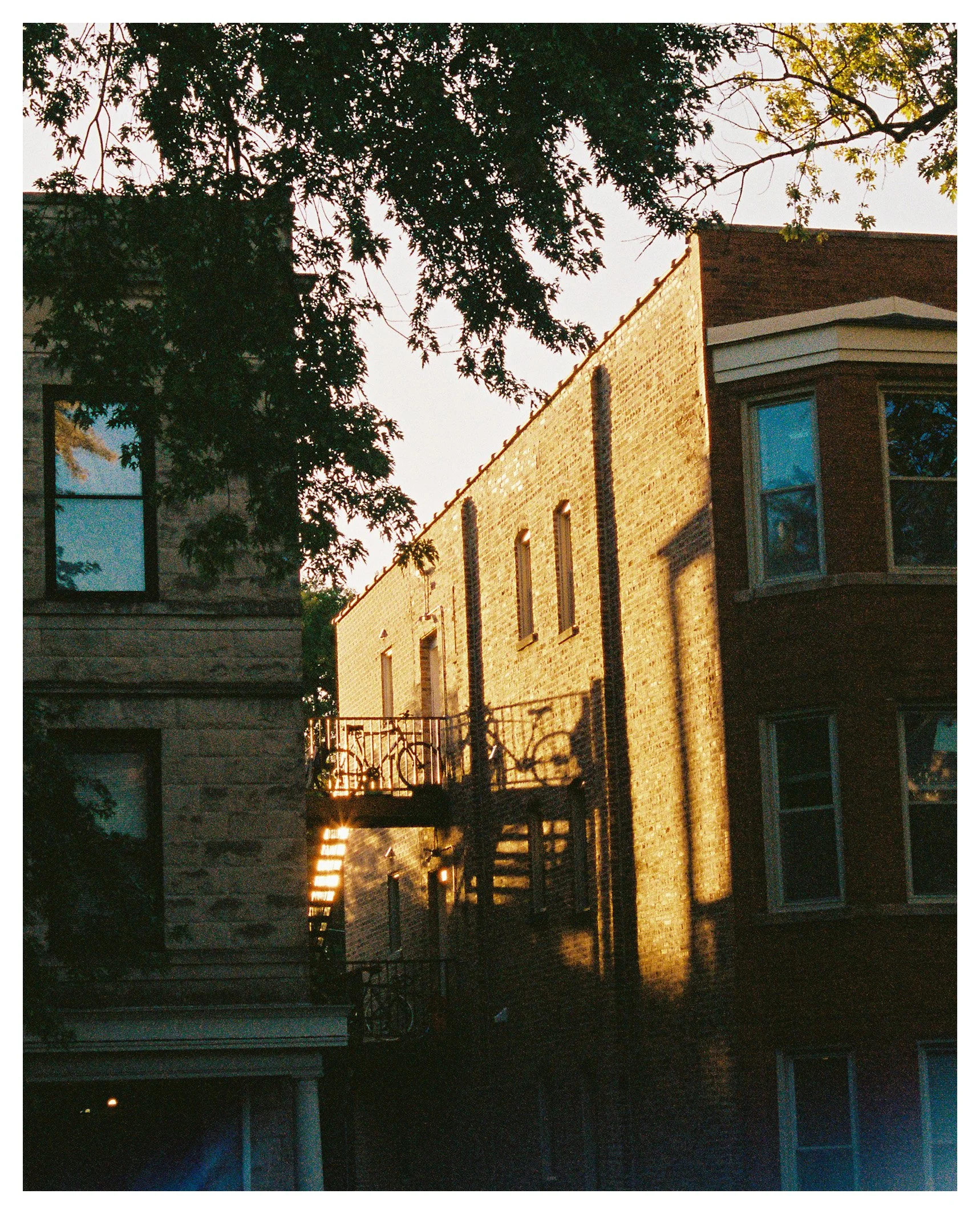 Sunset casting shadows on a brick apartment building with fire escape stairs, surrounded by trees.