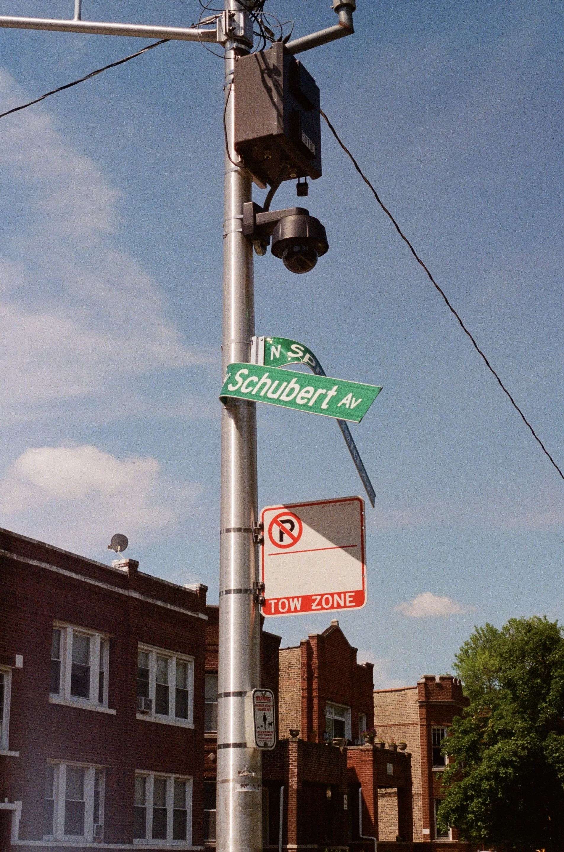 Street pole with street signs, traffic camera, and no parking and tow zone signs, with brick buildings and trees in the background under a blue sky.
