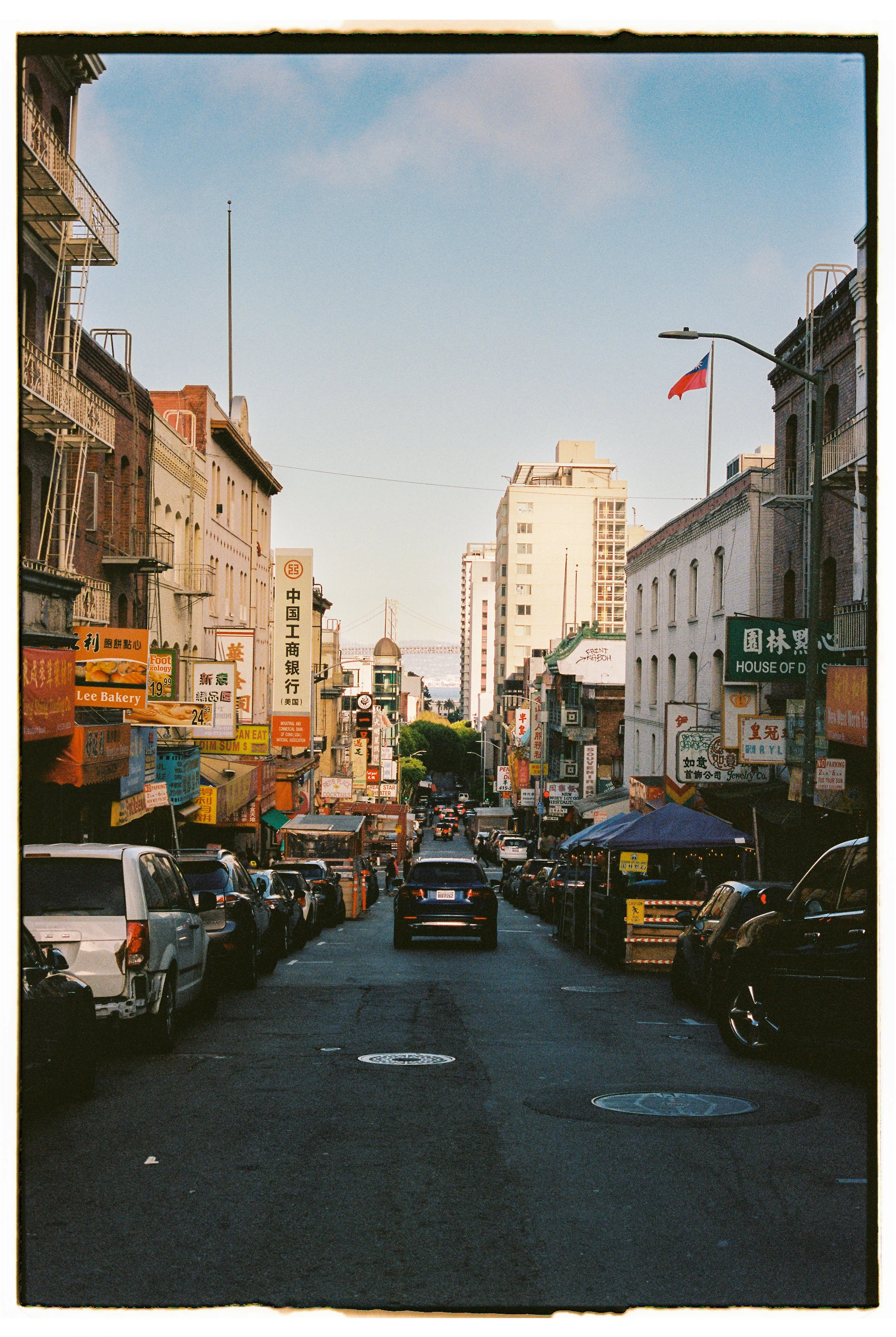 A city street lined with parked cars and storefronts with signs in Chinese theater-themed characters. Buildings in the background lead to a bridge over water under a clear sky.