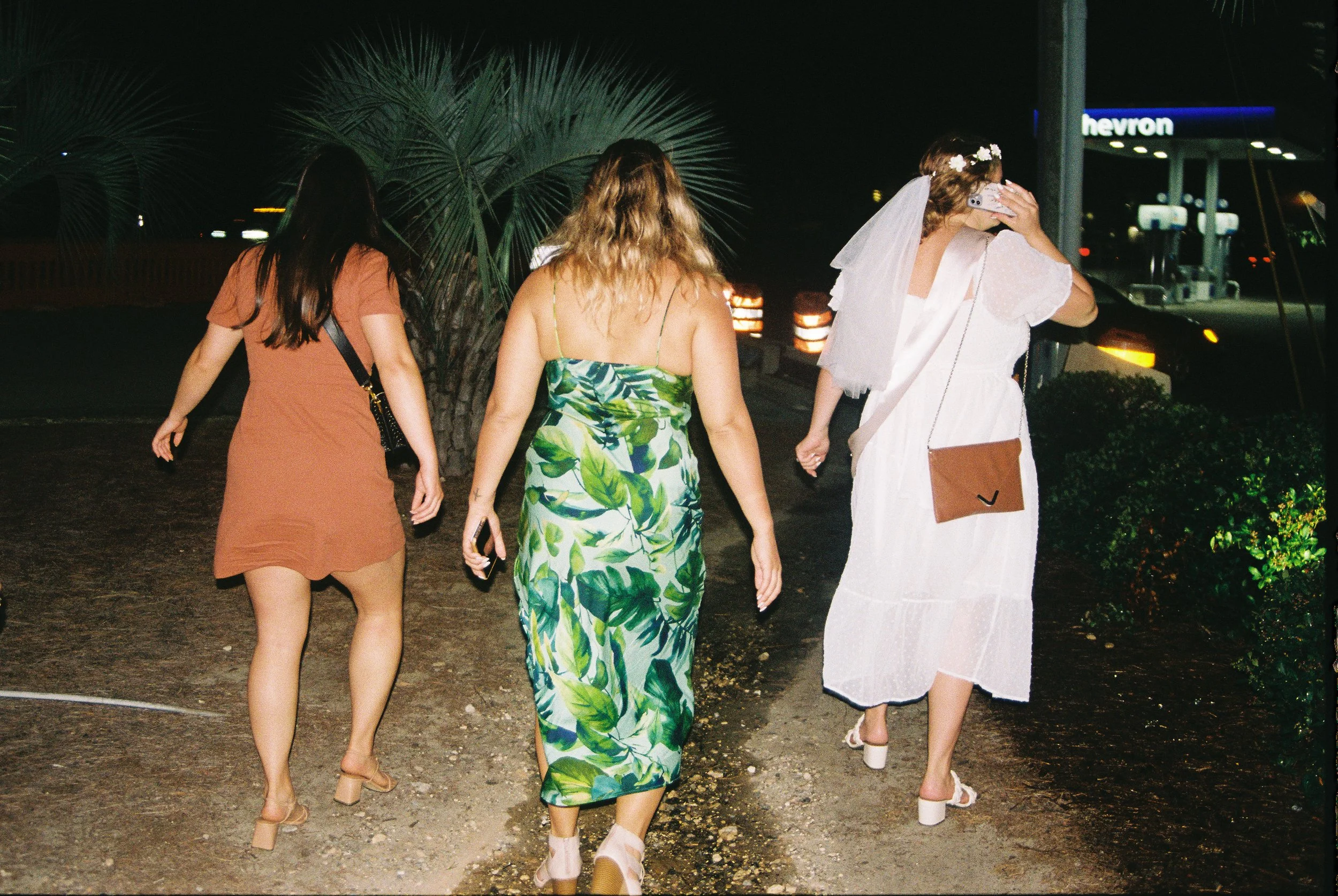 Three women walking away on a dirt path at night, with a Chevron gas station visible in the background, one wearing a brown dress, another in a green leafy dress, and the third in a white dress with a veil.