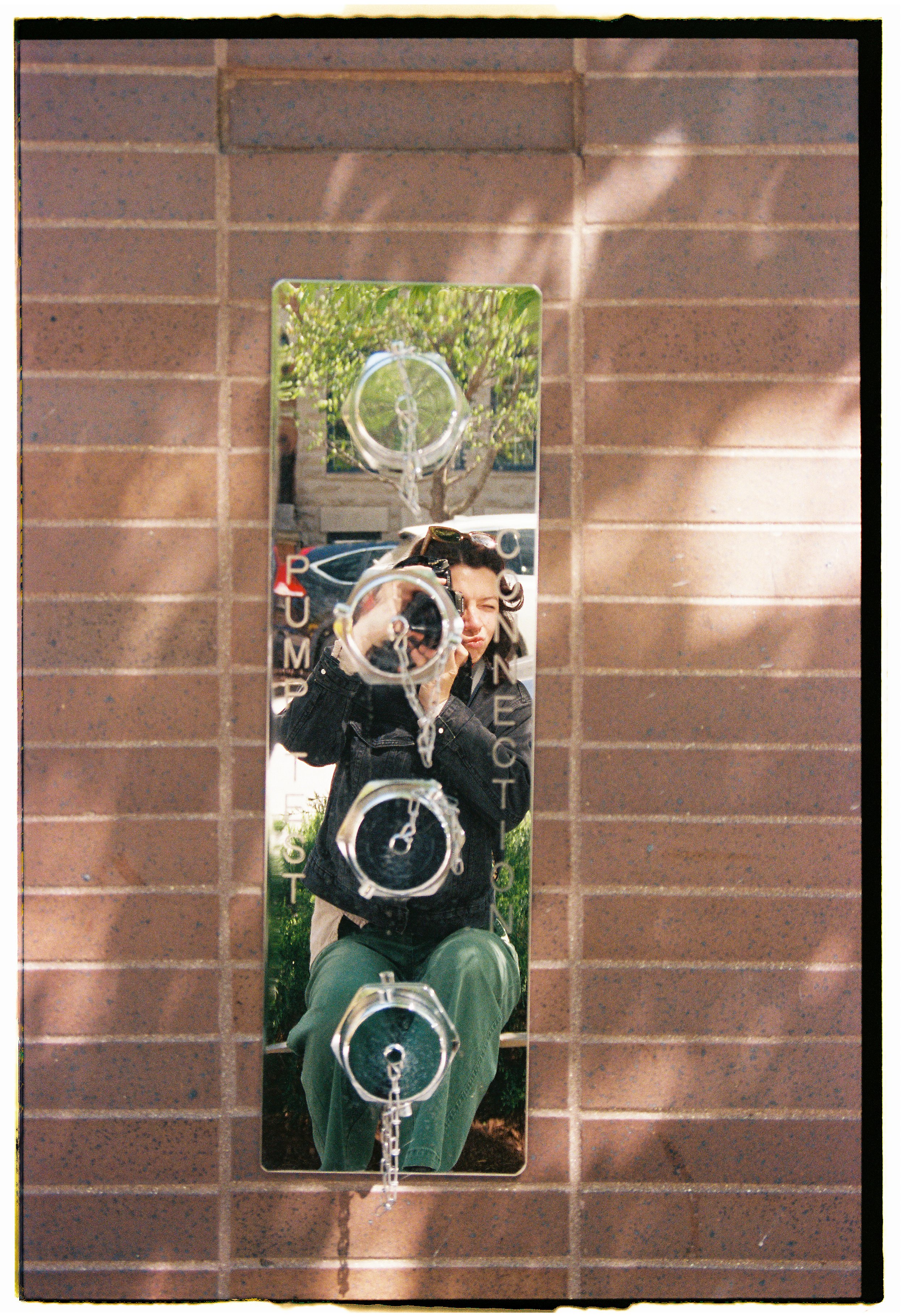 A woman taking a photo with a camera is reflected in a small vertical mirror mounted on a brick wall. The mirror has the words 'TRUMPET' and other words written on it, and there are chain loops hanging from the mirror's top.