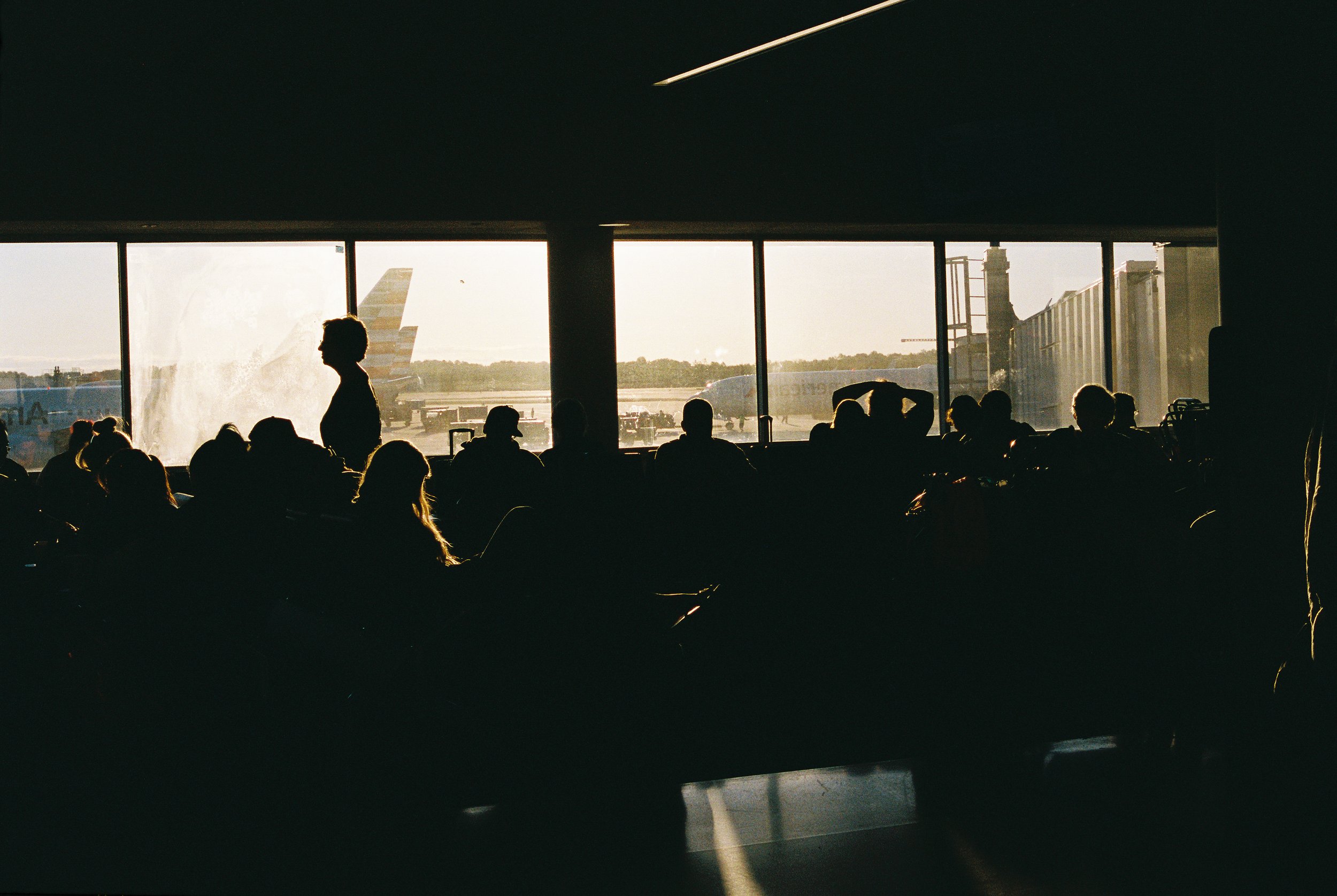 Silhouettes of travelers seated in an airport waiting area, with large windows showing airplanes and airport tarmac outside during sunset or sunrise.